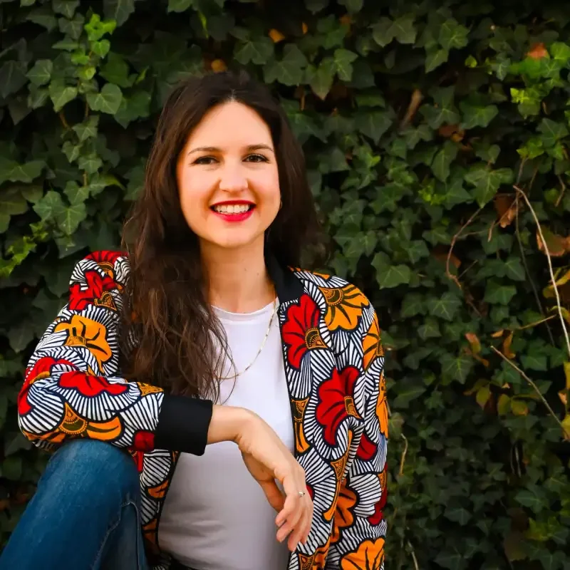 Woman with long brown hair and red lipstick sitting in front of green ivy wall, wearing a floral patterned jacket and white shirt.