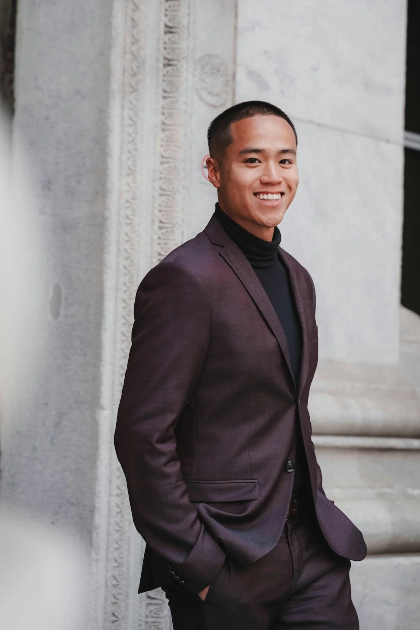 A young man in a dark suit and black turtleneck, smiling, with short hair, standing against a stone building.
