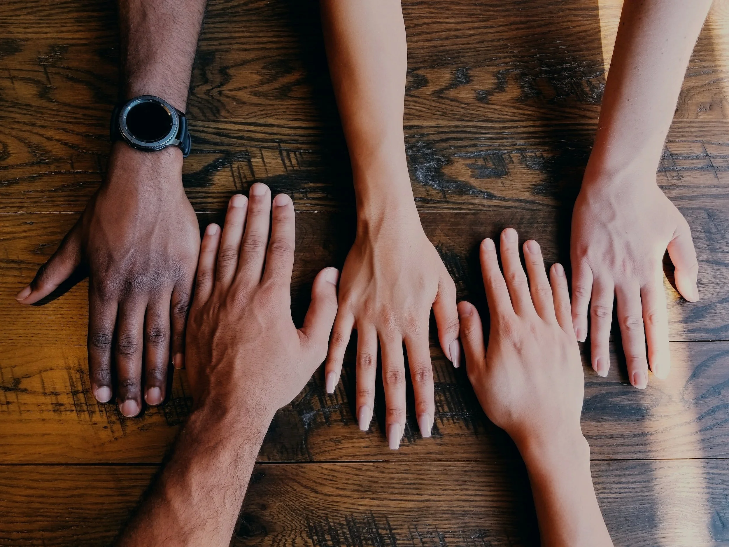 Multiple hands of different skin tones stacked on a wooden table.