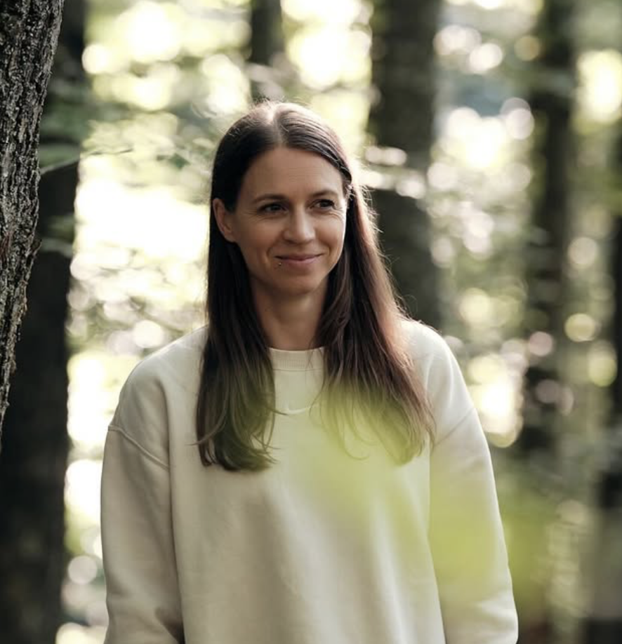 A woman with dark hair and a friendly smile, wearing a sleeveless top, sitting indoors at a table with blurred background