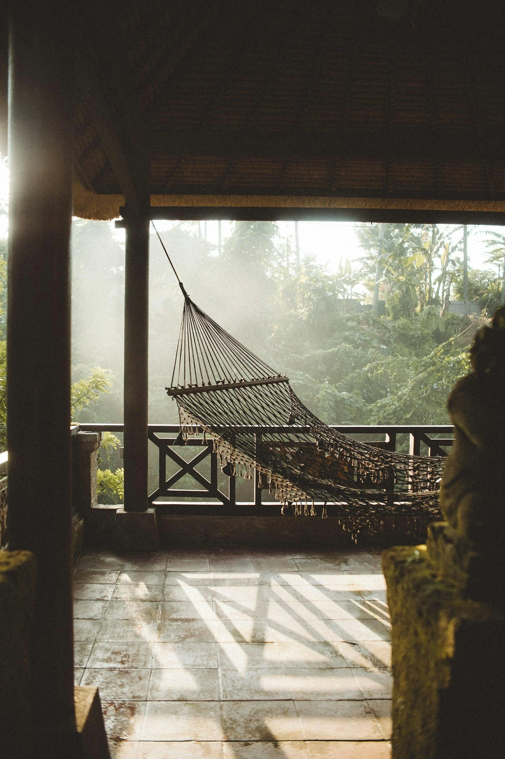 A hammock hanging on a balcony overlooking a lush, green tropical garden, with sunlight filtering through trees in the background.