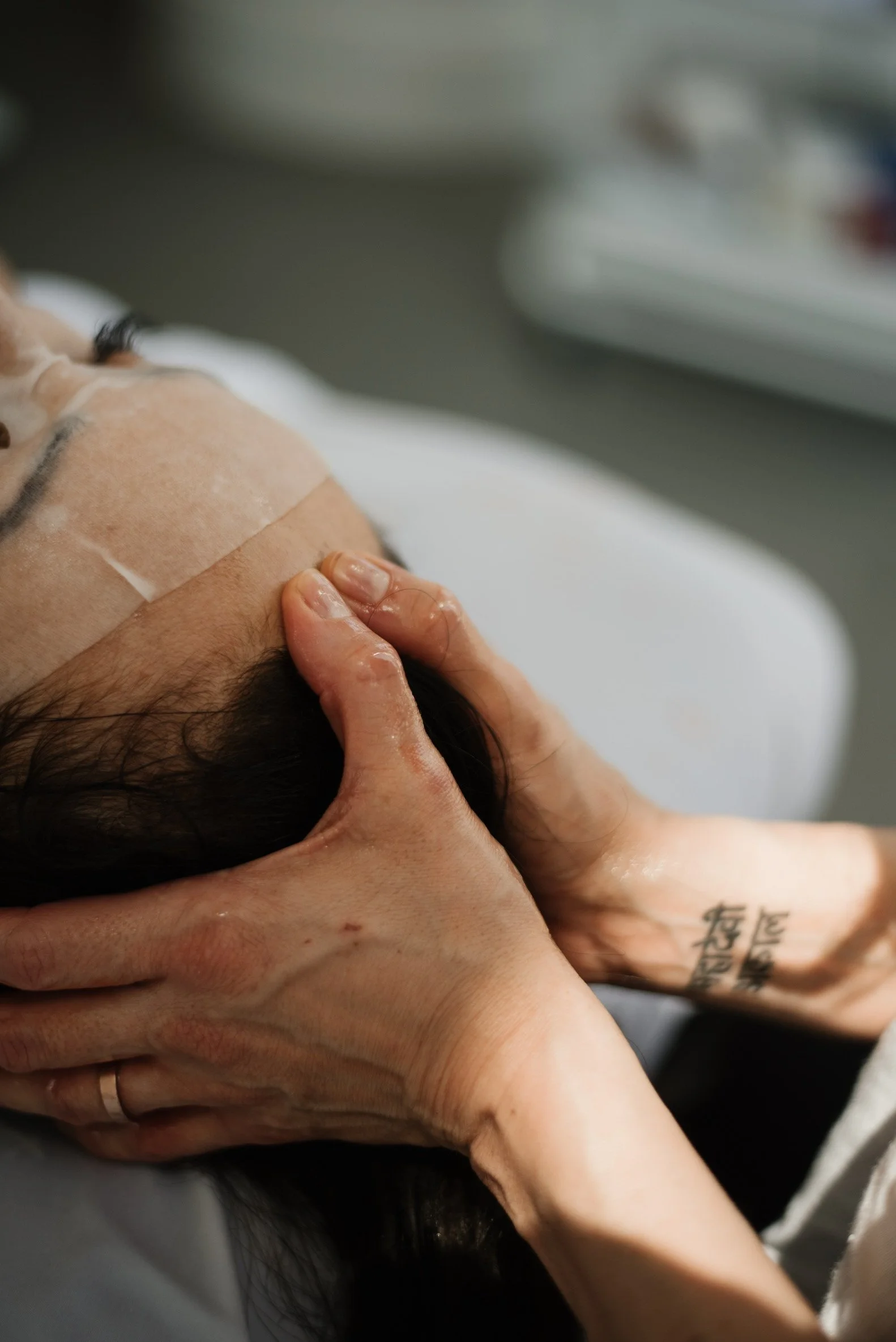 A person receiving a head massage or treatment, with hands gently holding their head. The person has dark hair and is lying down.