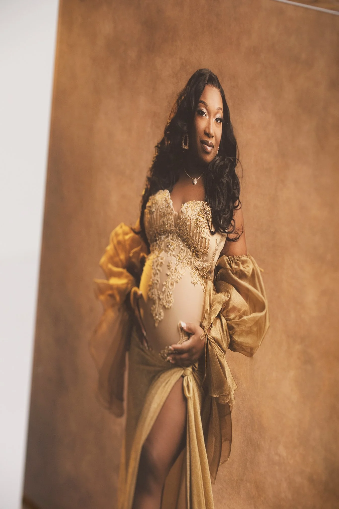 Portrait of a woman with long curly black hair, wearing a gold-colored dress with intricate embroidery, jewelry, and puffed sleeves, standing against a plain brown background.