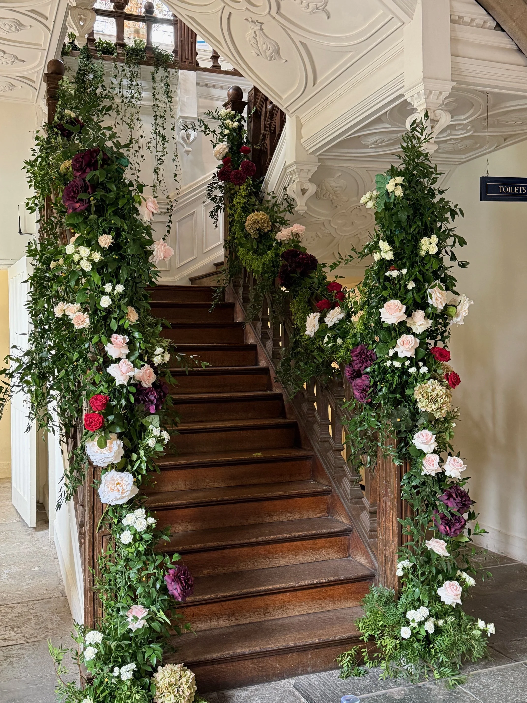 A staircase decorated with a variety of colorful flowers and green foliage on both sides, leading up to a second floor with wooden railings and a decorative ceiling.