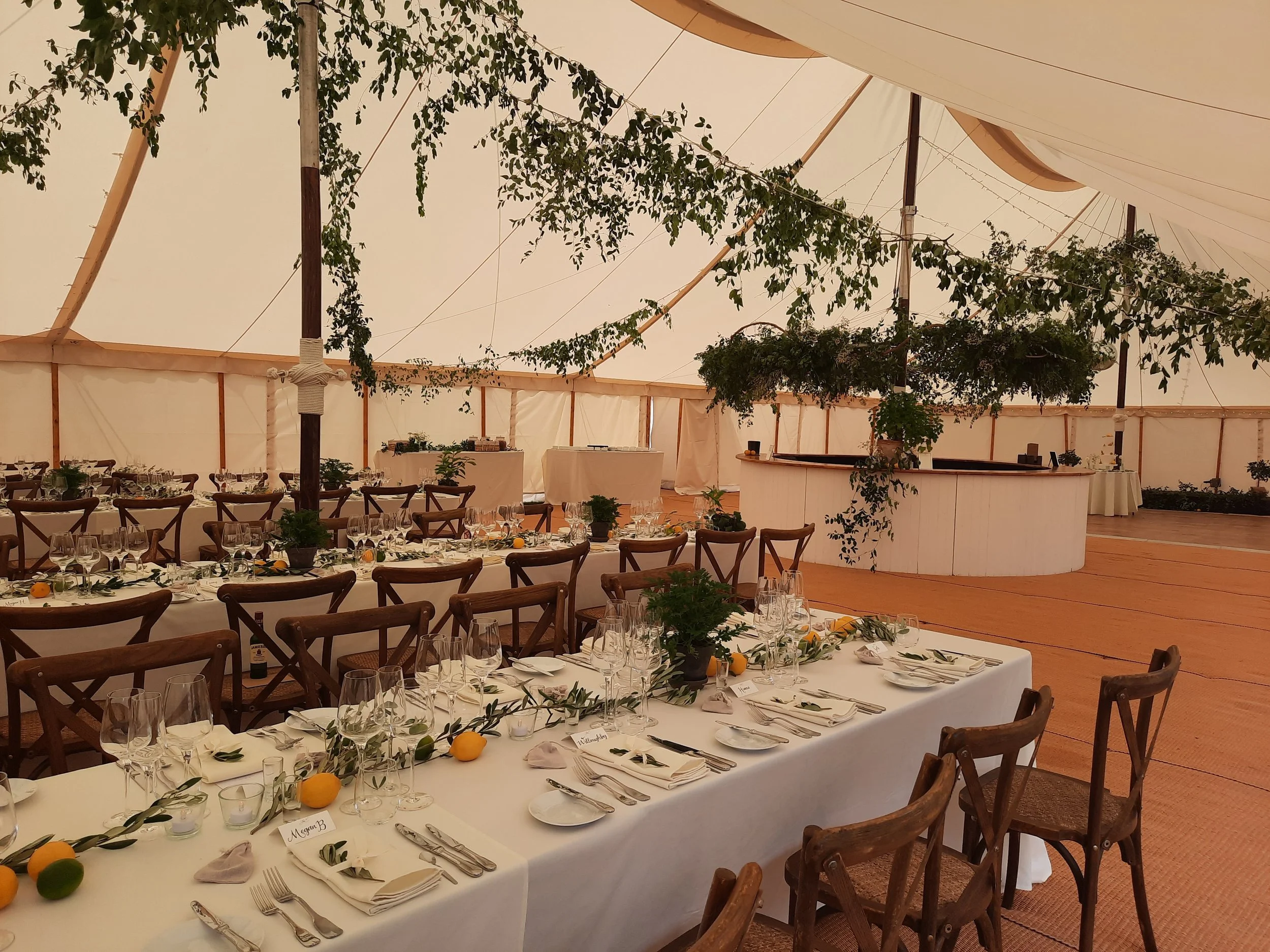 Elegant indoor event tent with round and rectangular tables set for a formal dinner, decorated with greenery, lemons, and tableware, with a wooden floor and white drapes.