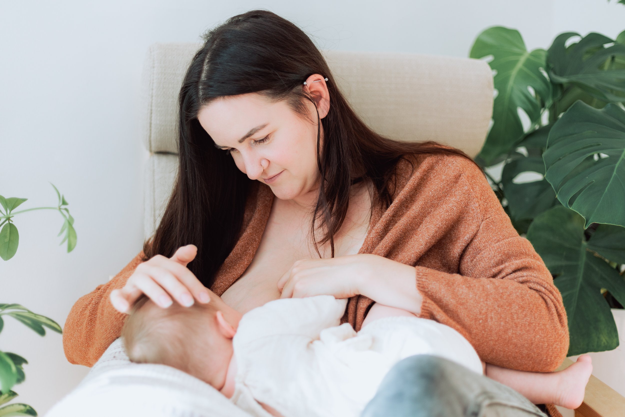 Woman breastfeeding a baby seated on a beige chair, surrounded by green plants.