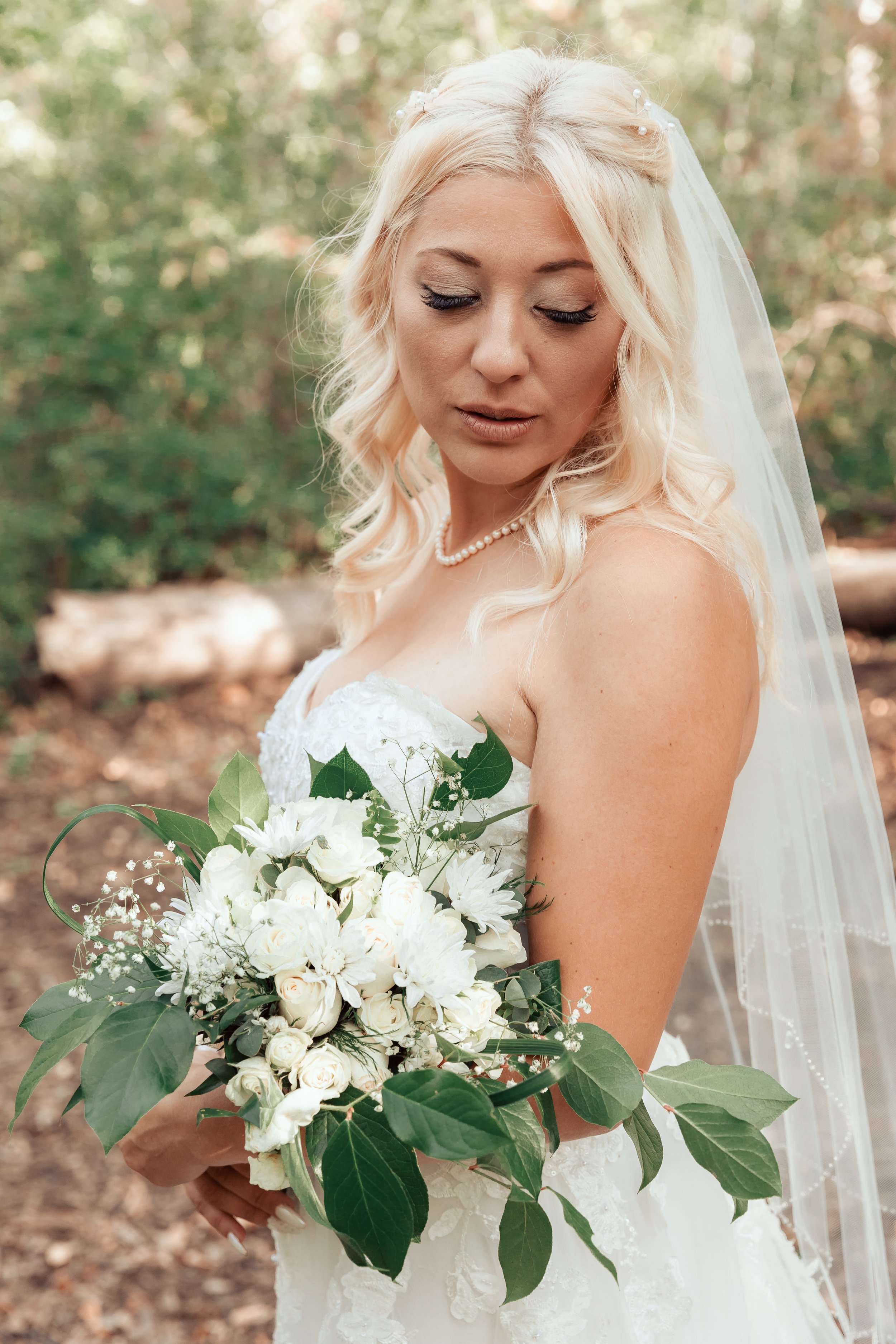 Bride in white dress holding a bouquet of white flowers and green leaves, standing outdoors.