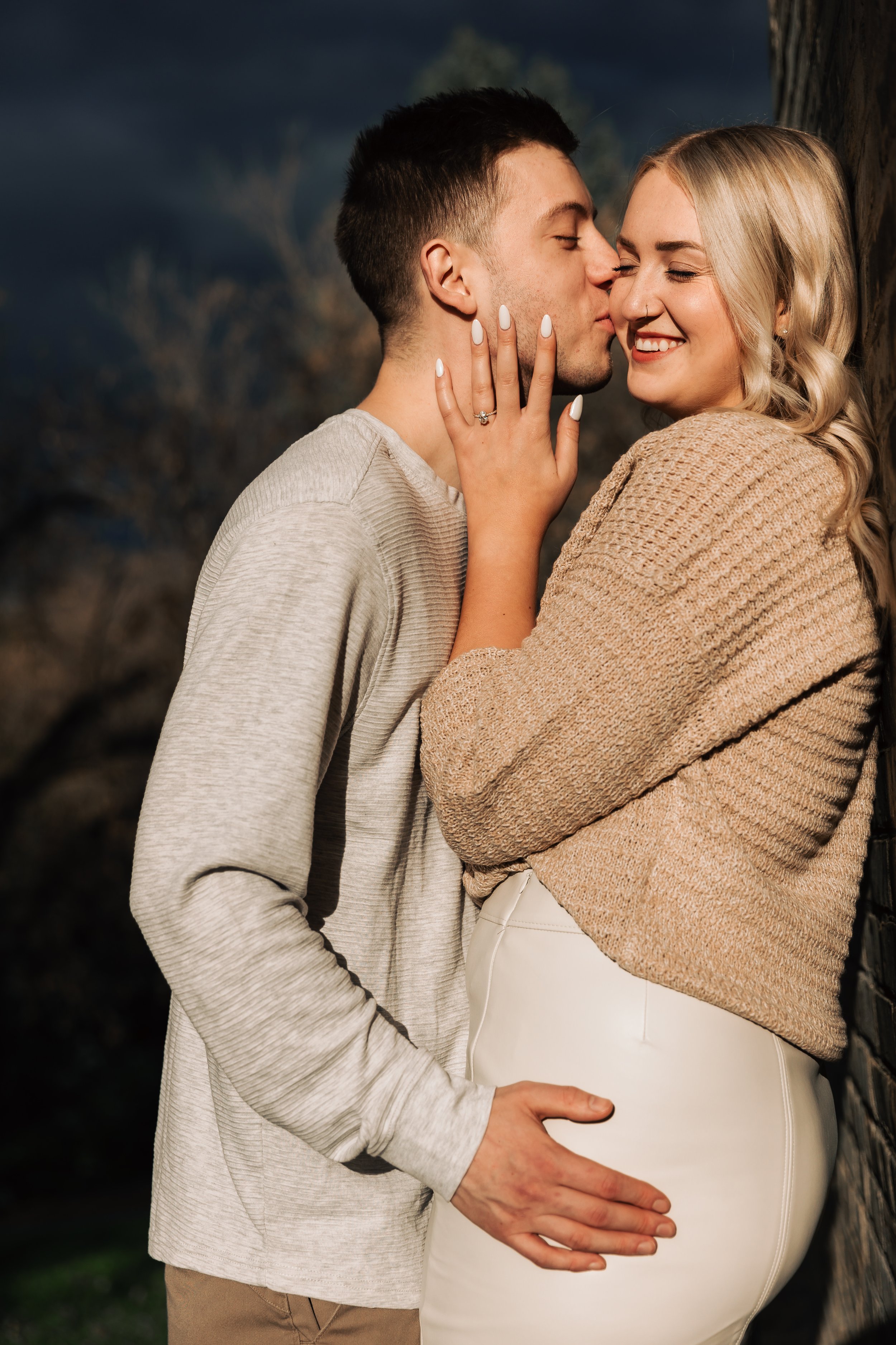 A couple embracing, the woman smiling with engagement ring visible, outdoors with dark sky background.