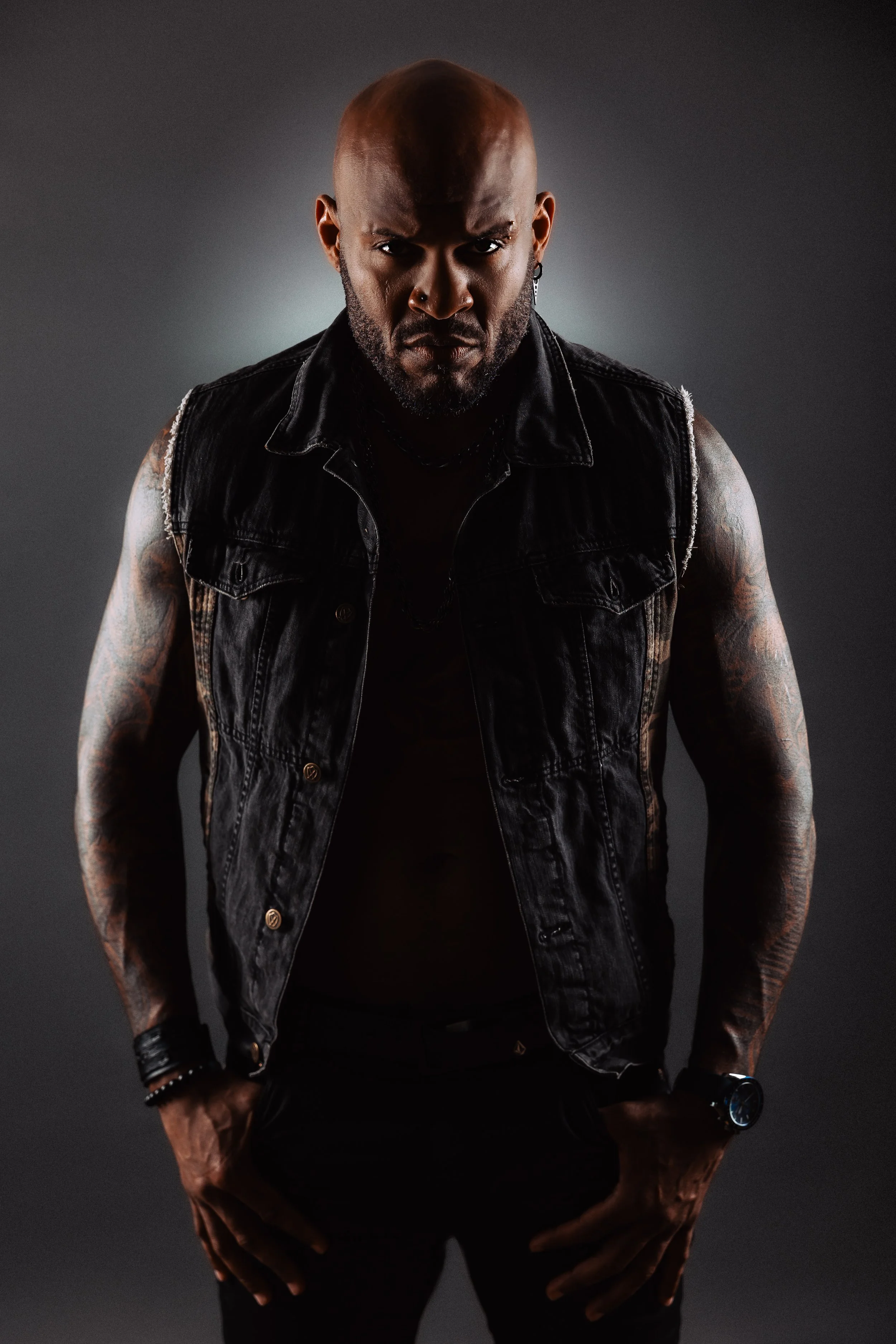 A bald man with a beard, wearing a sleeveless black denim vest, black shirt, tattoos on arms, and accessories including earrings, a smartwatch, and bracelets, standing against a dark background.