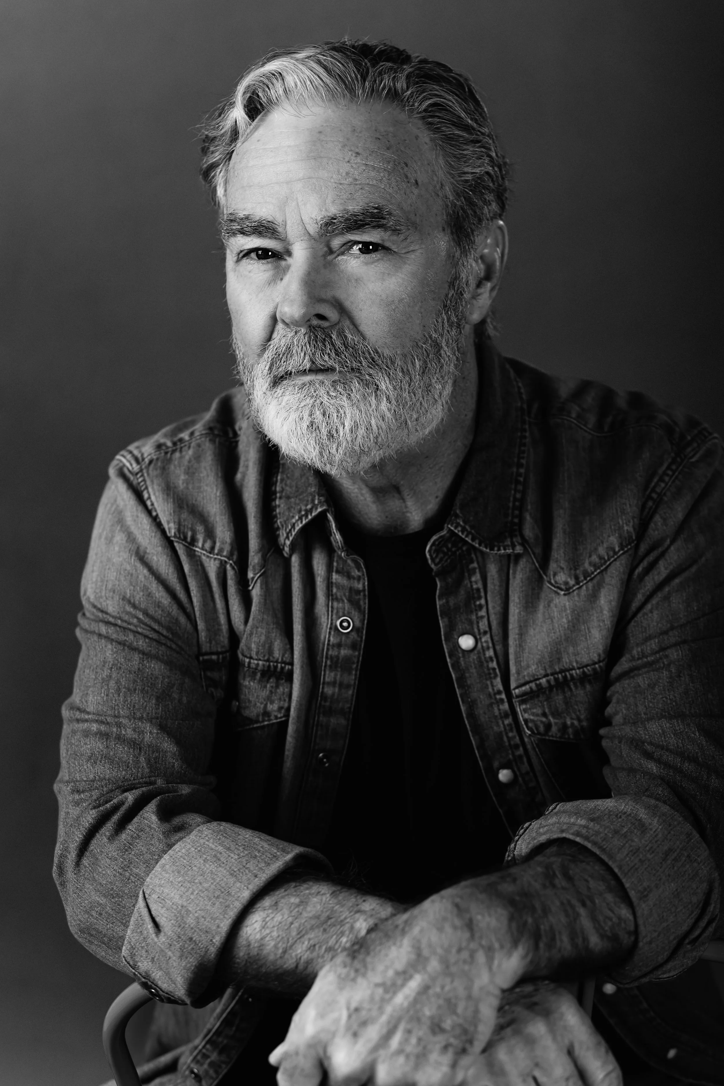 Black and white portrait of an elderly man with a beard and hair, wearing a denim shirt, looking directly at the camera with a serious expression.