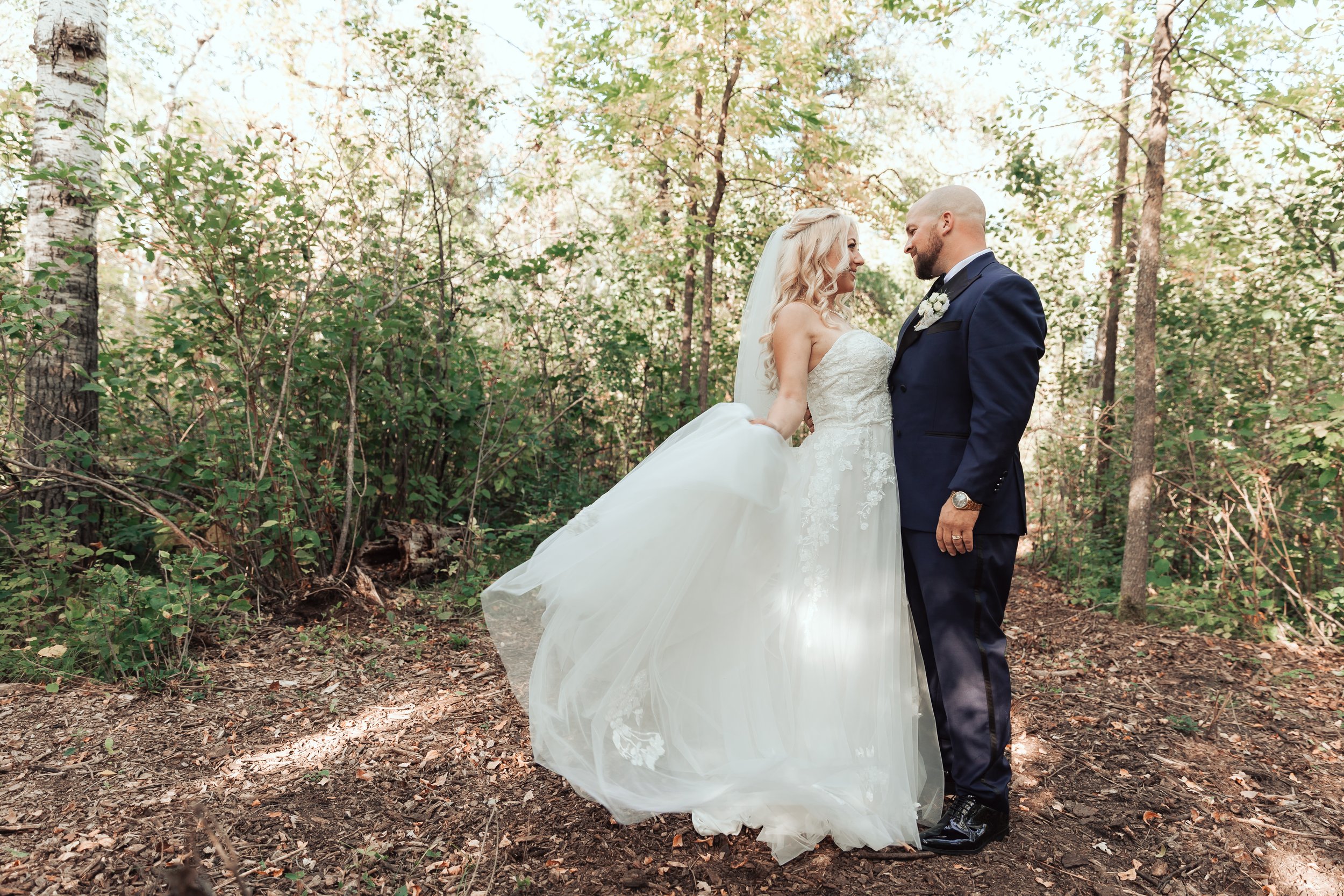 Bride and groom standing in forest, bride holding dress, groom in dark suit, surrounded by trees.