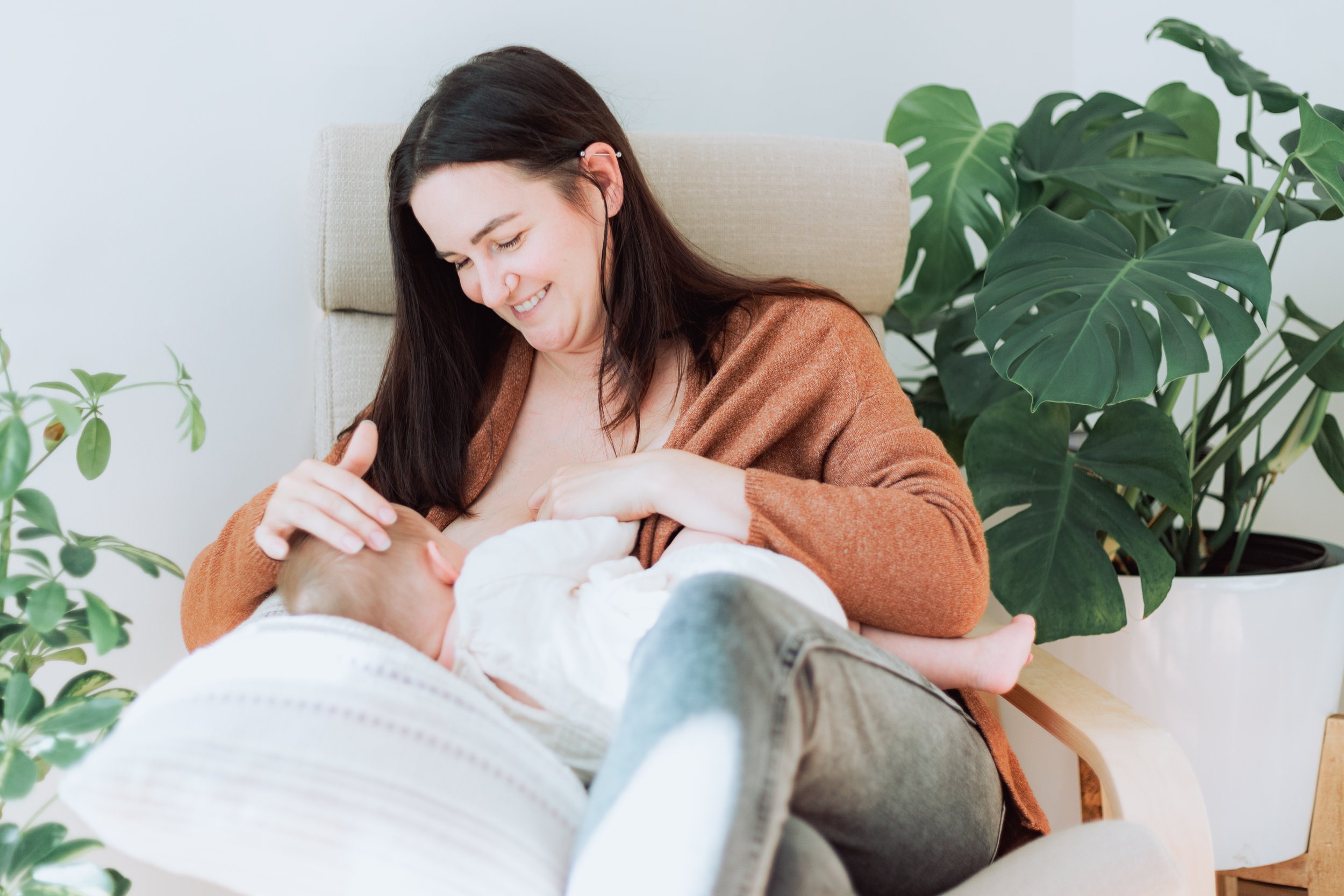 A woman breastfeeding her baby while sitting in a chair, surrounded by green plants.