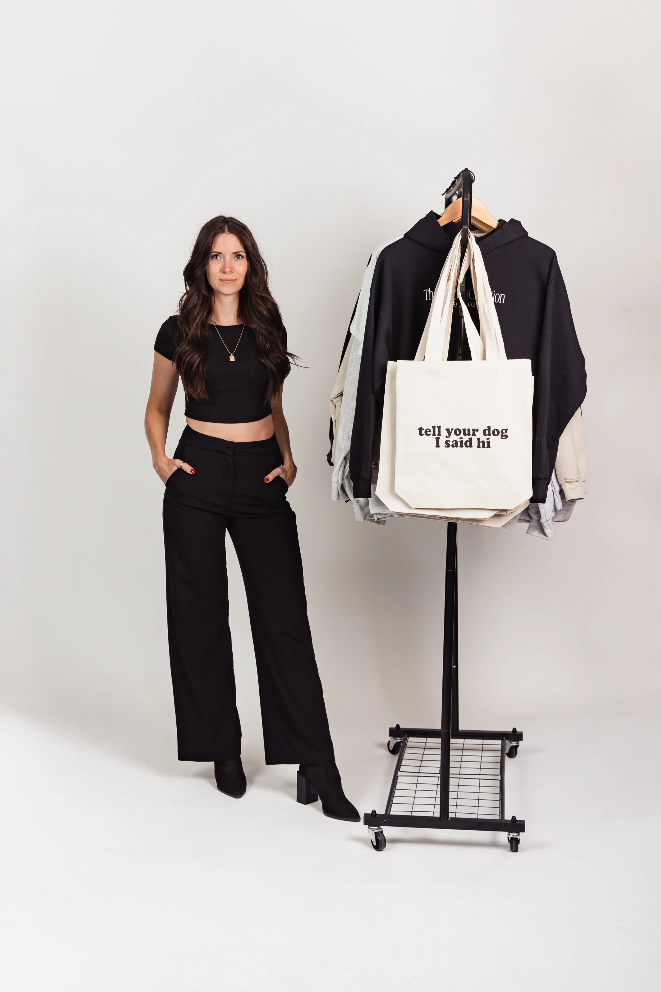A woman standing next to a clothing rack with hoodies and tote bags in a studio setting.
