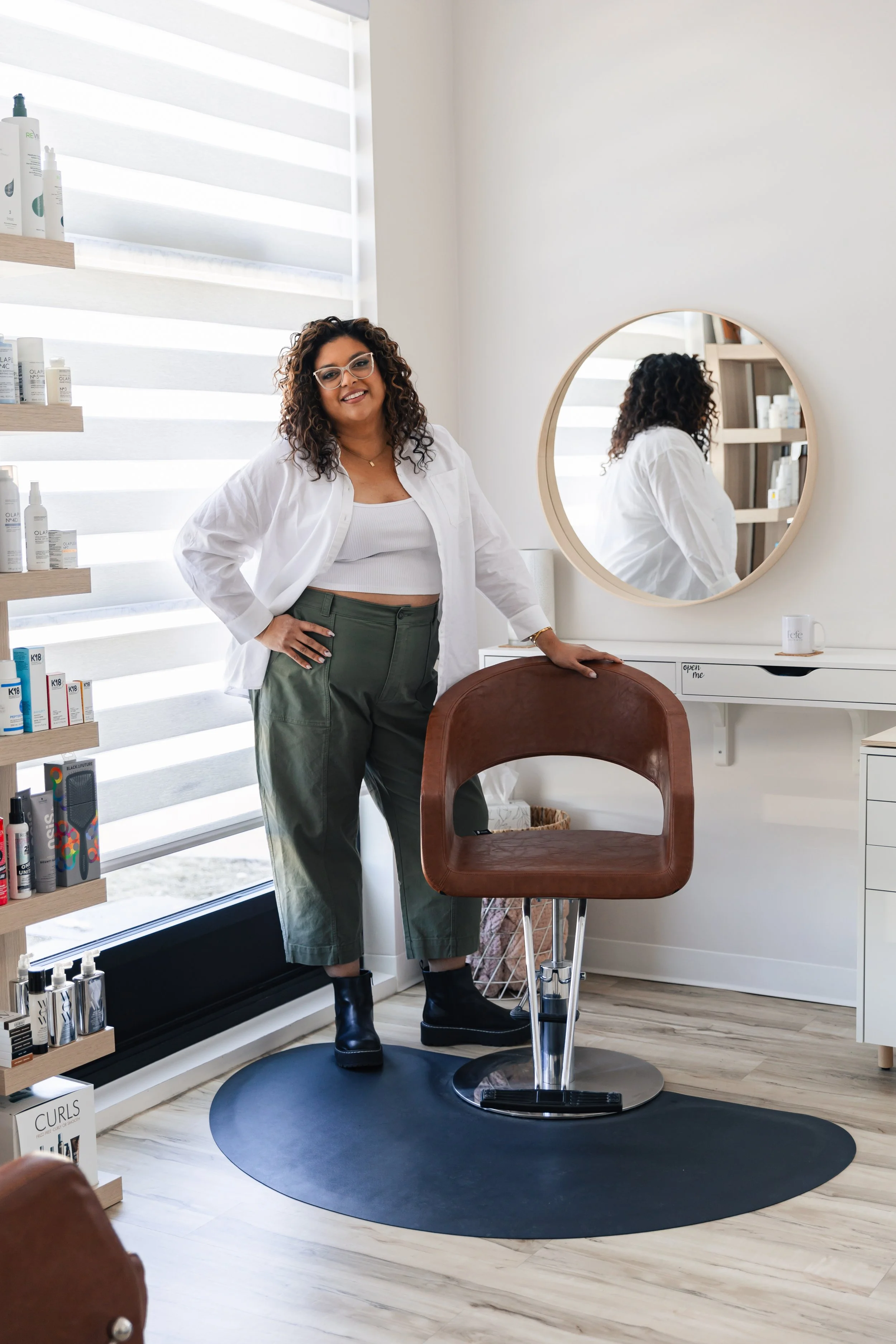 A woman standing in a hair salon next to a chair, with shelves of hair products behind her, a mirror on the wall, and a desk with a mug. She has curly hair, glasses, and is wearing a white shirt, green pants, and black boots.
