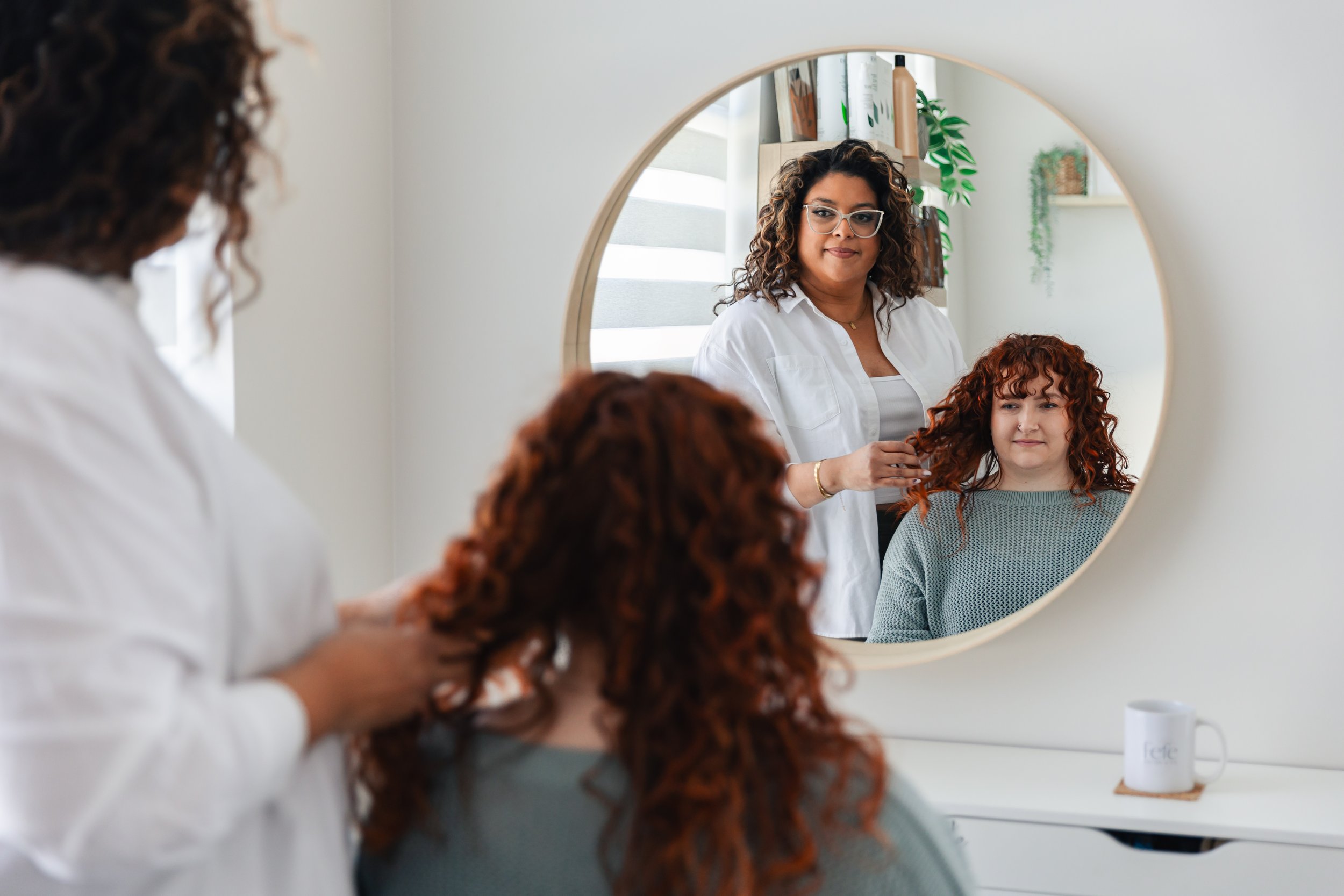 A hairstylist with glasses and curly hair is styling a red-haired woman's curly hair in front of a round mirror in a bright, modern room with plants and books.