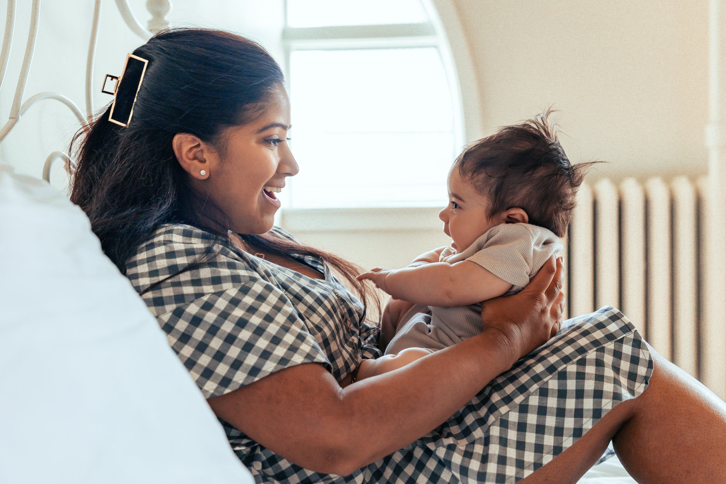 A woman holding a baby on a bed, both smiling at each other, with natural light coming from a window in the background.