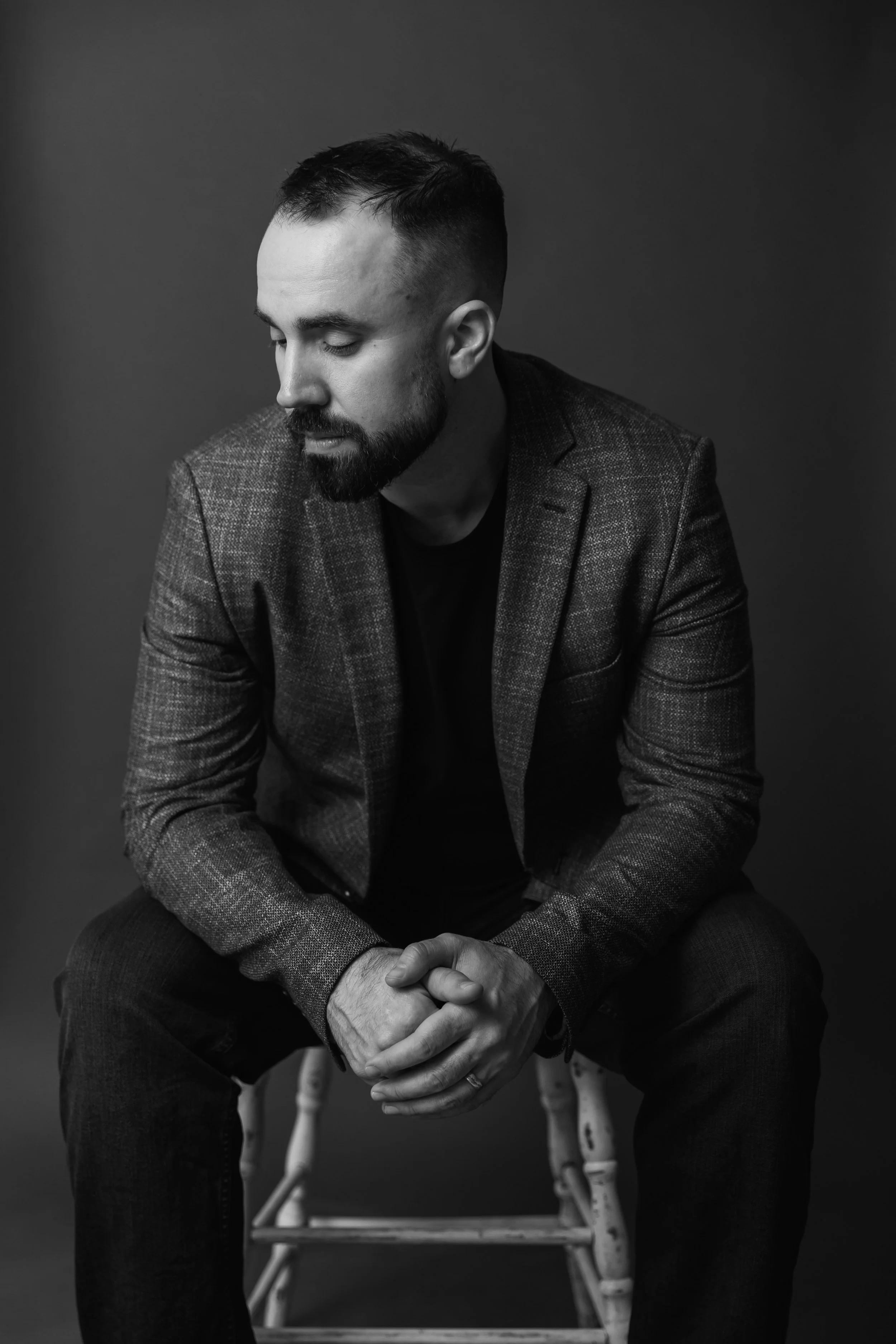A black and white photo of a man with a beard and short hair, sitting on a rustic wooden chair, looking down with hands clasped, dressed in a blazer and dark shirt against a plain background.