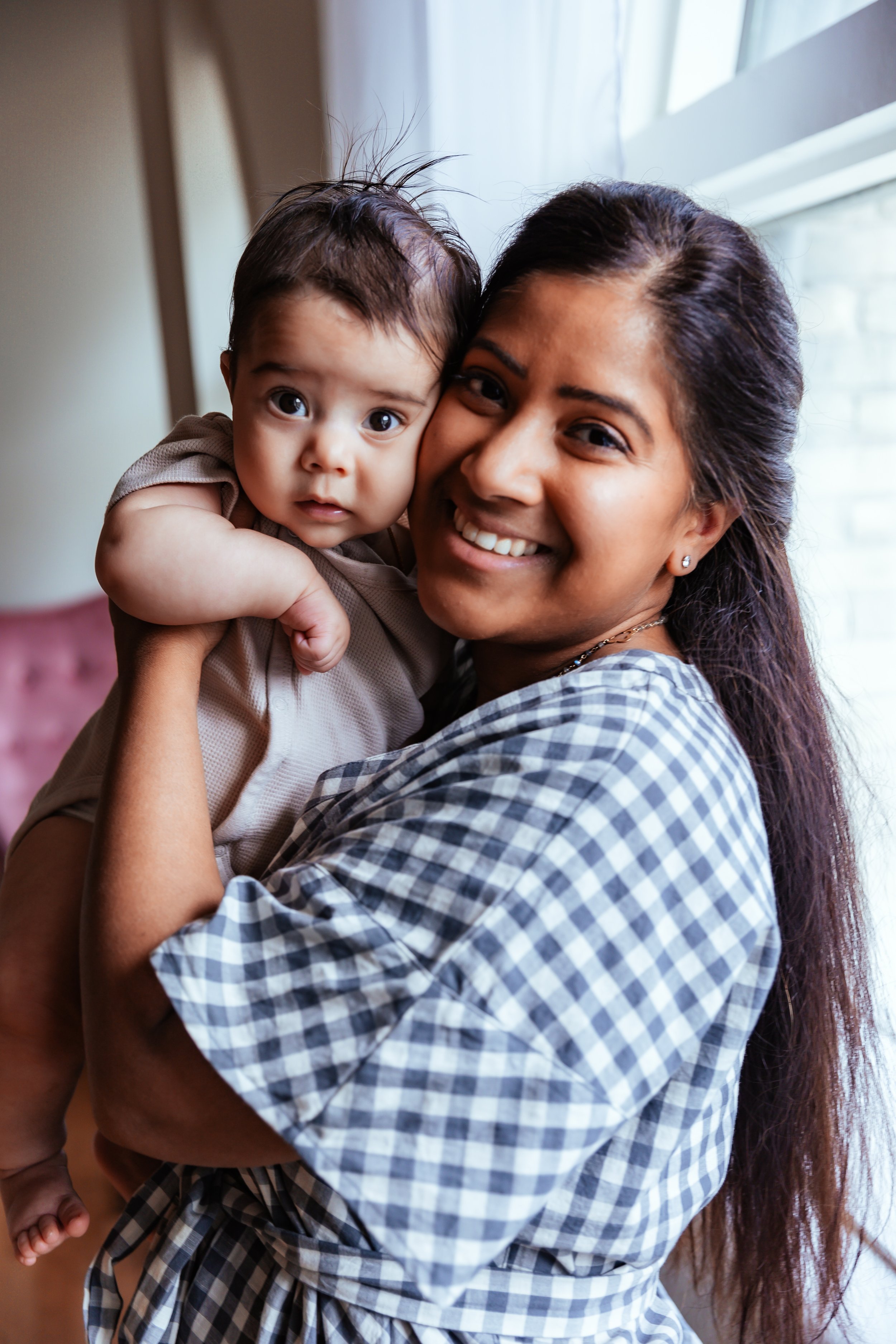 A woman holding a baby near a window, both smiling. The woman is wearing a checkered shirt.