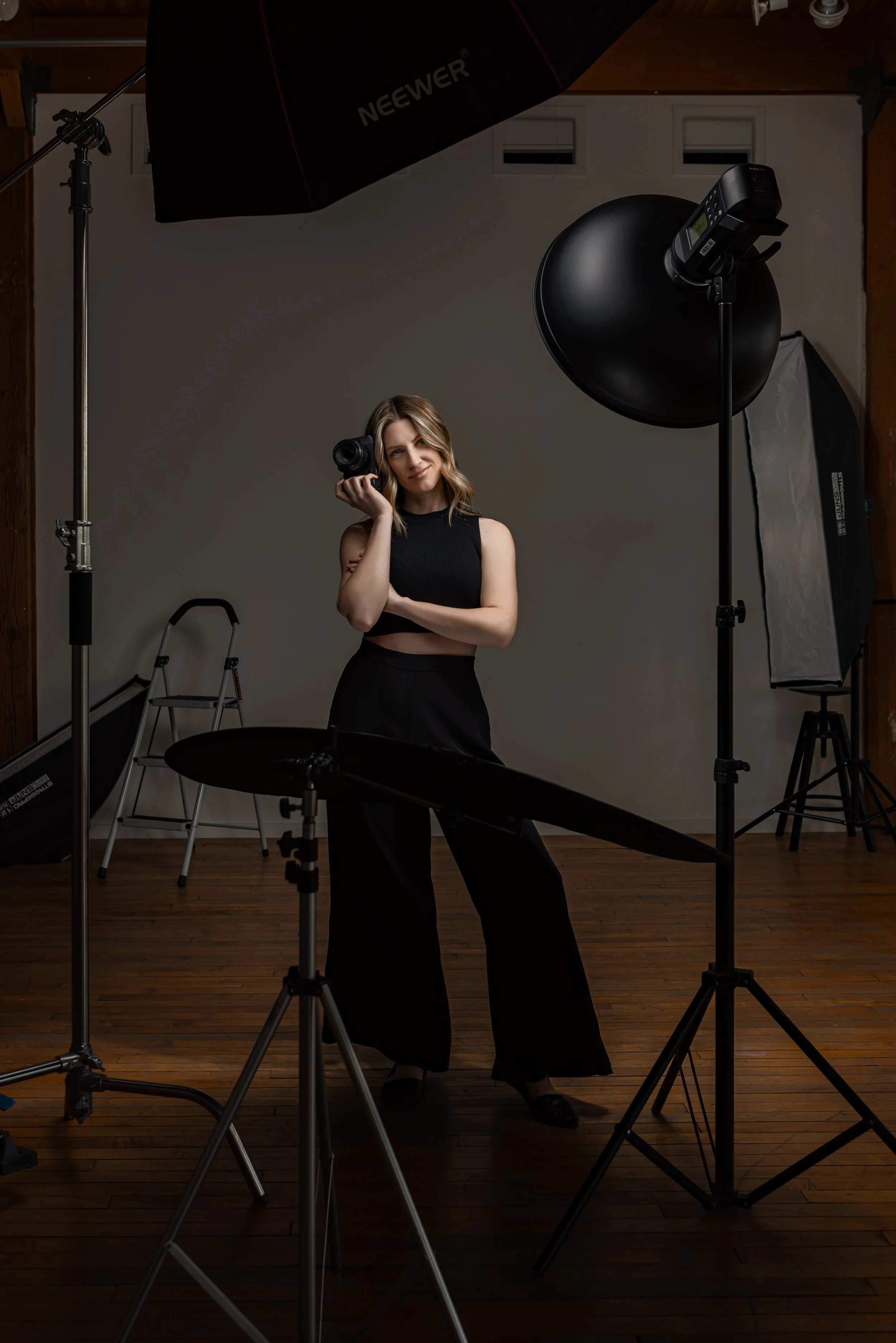 A woman in a photography studio holding a camera, surrounded by professional lighting and photography equipment.