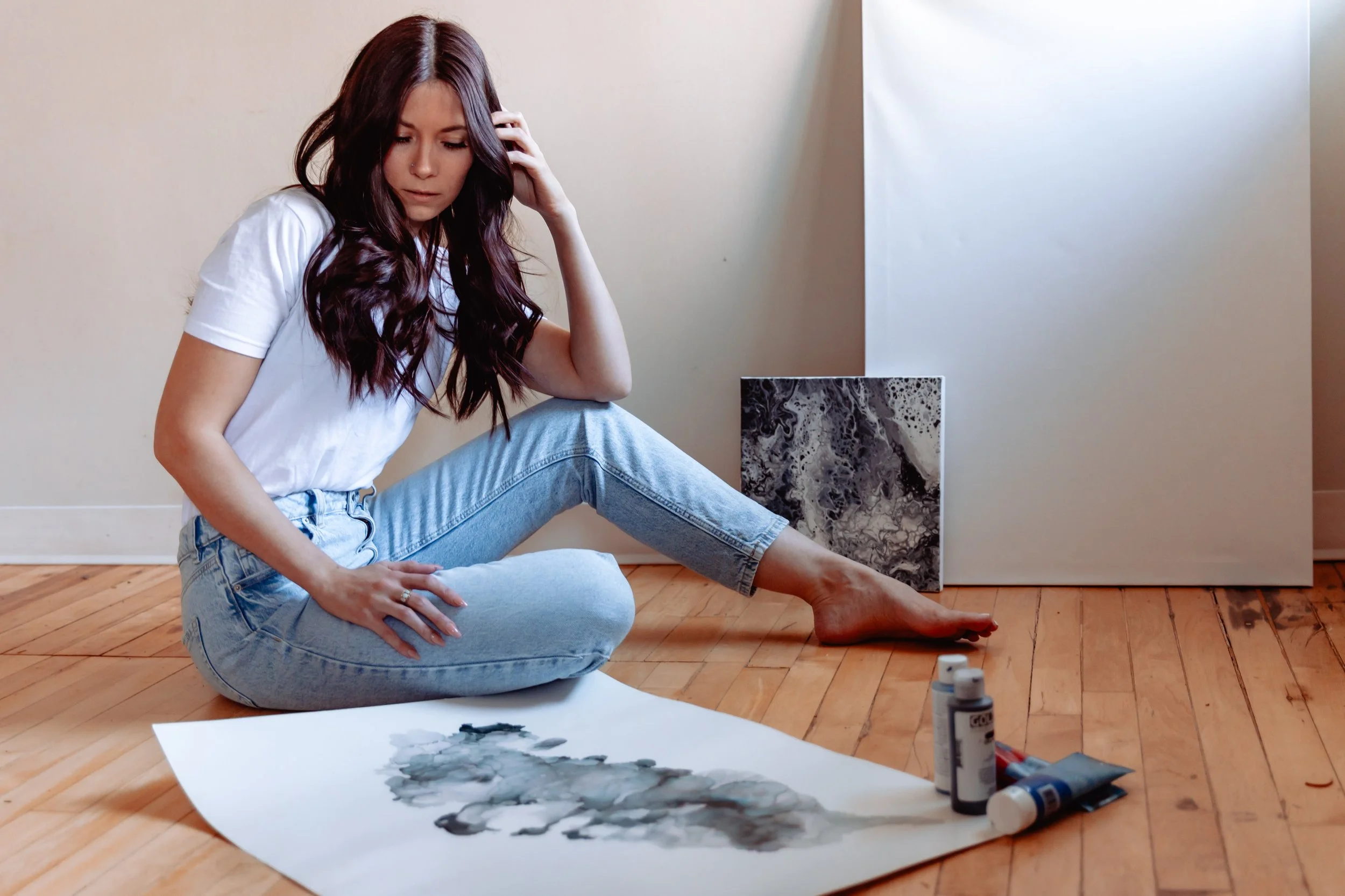 A woman with long brown hair, wearing a white t-shirt and light blue jeans, sitting on a wooden floor and looking at a watercolor painting on paper in front of her. There are bottles of paint or ink nearby, and a large abstract black and white painti