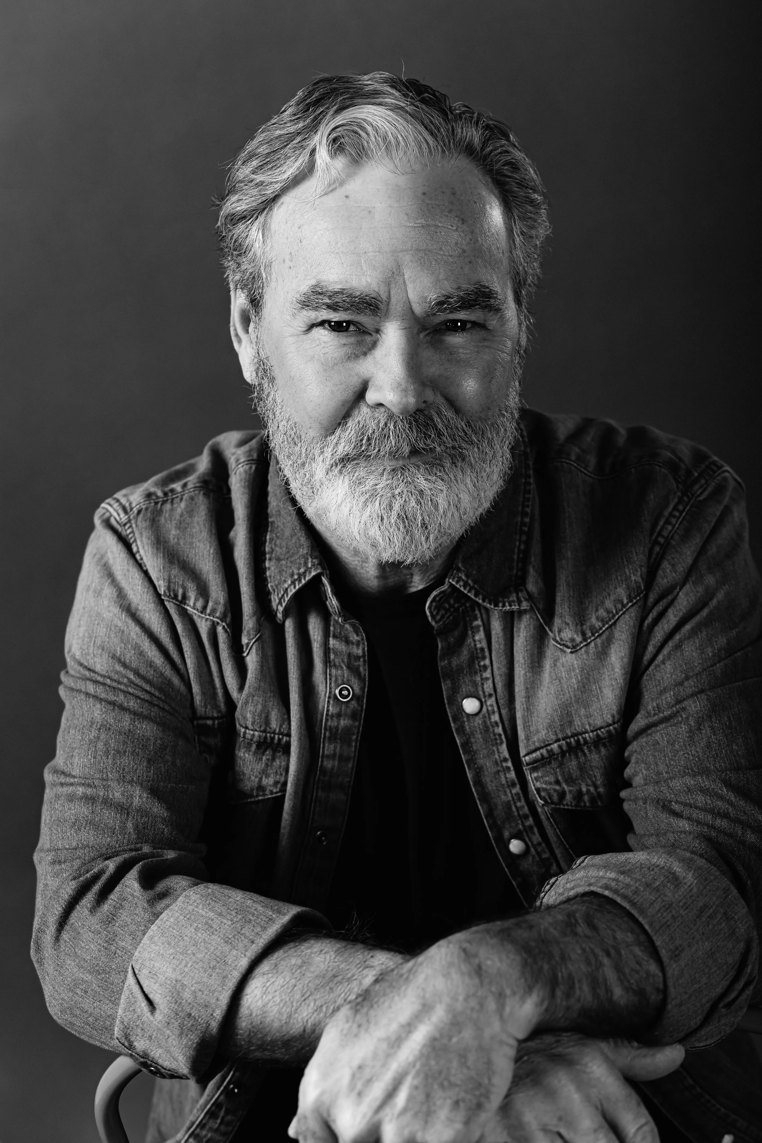 Black and white portrait of an older man with a beard and mustache, wearing a denim jacket and resting his arm on a chair.