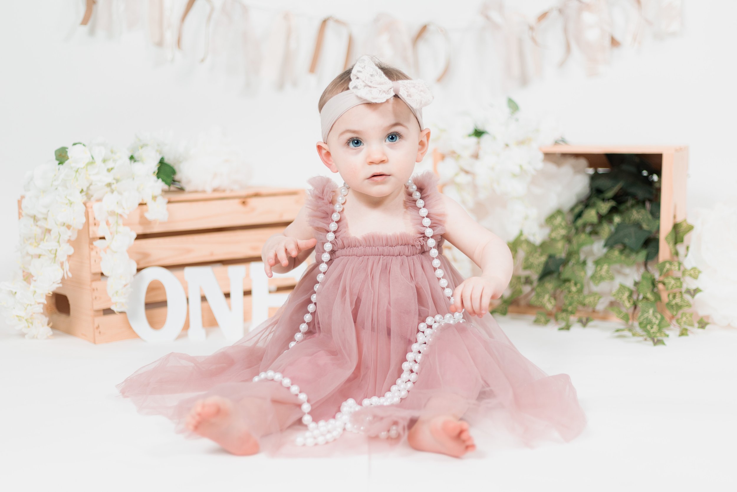 Baby in a pink dress sitting with a wooden crate, white flowers, and green leaves, holding a string of white pearls. A "ONE" sign indicates a first birthday celebration.