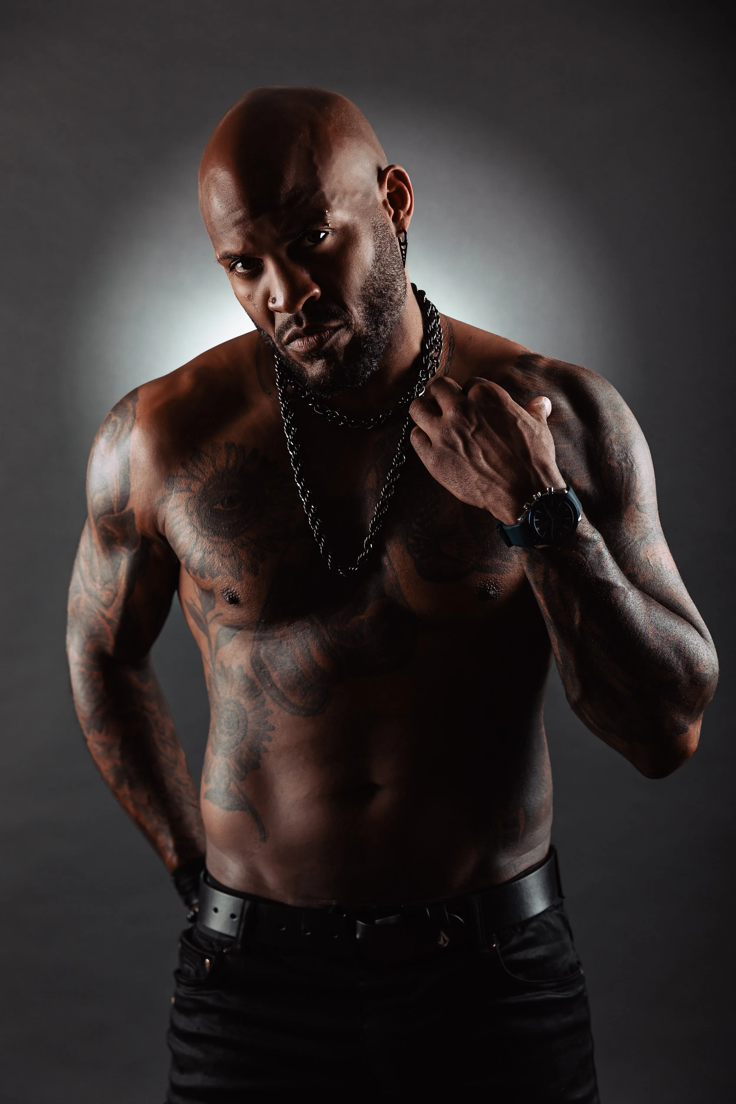 A muscular, tattooed man with a shaved head poses shirtless against a dark background, wearing chains, a black watch, and black pants.