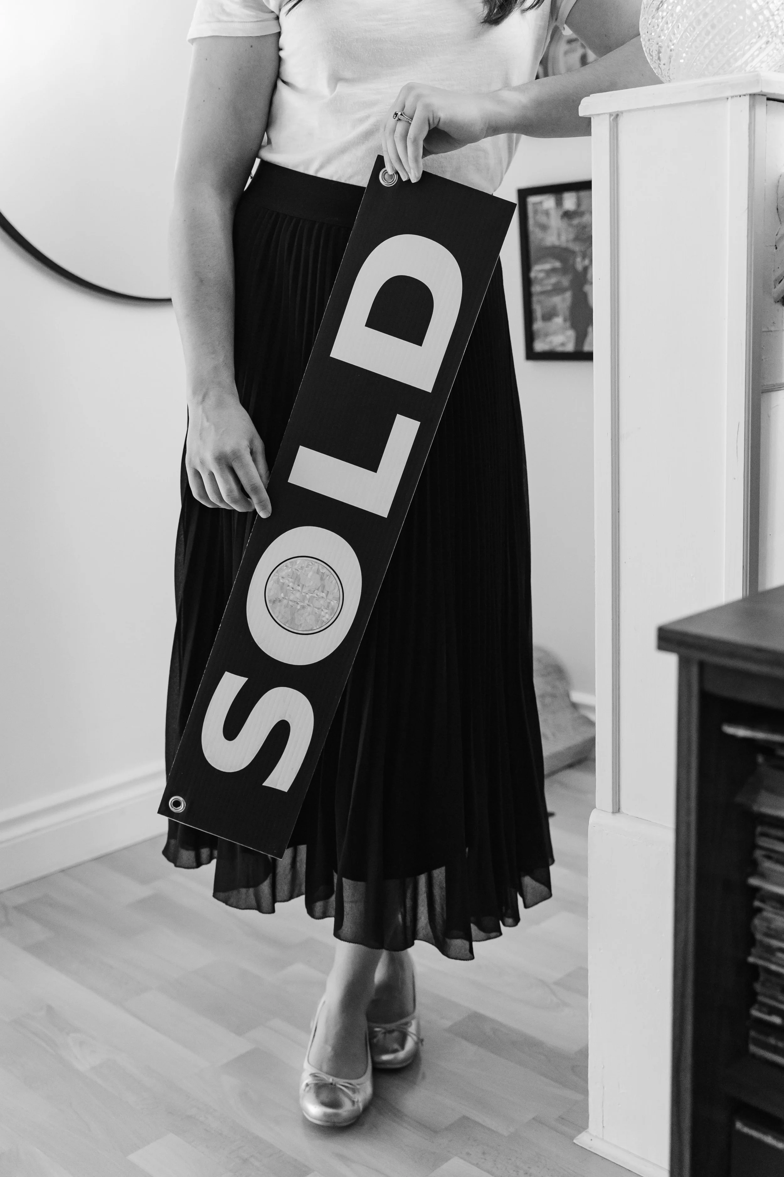 A woman holding a large sign that says 'SOLD' in black and white, standing in a room with hardwood floors and a small furniture piece.