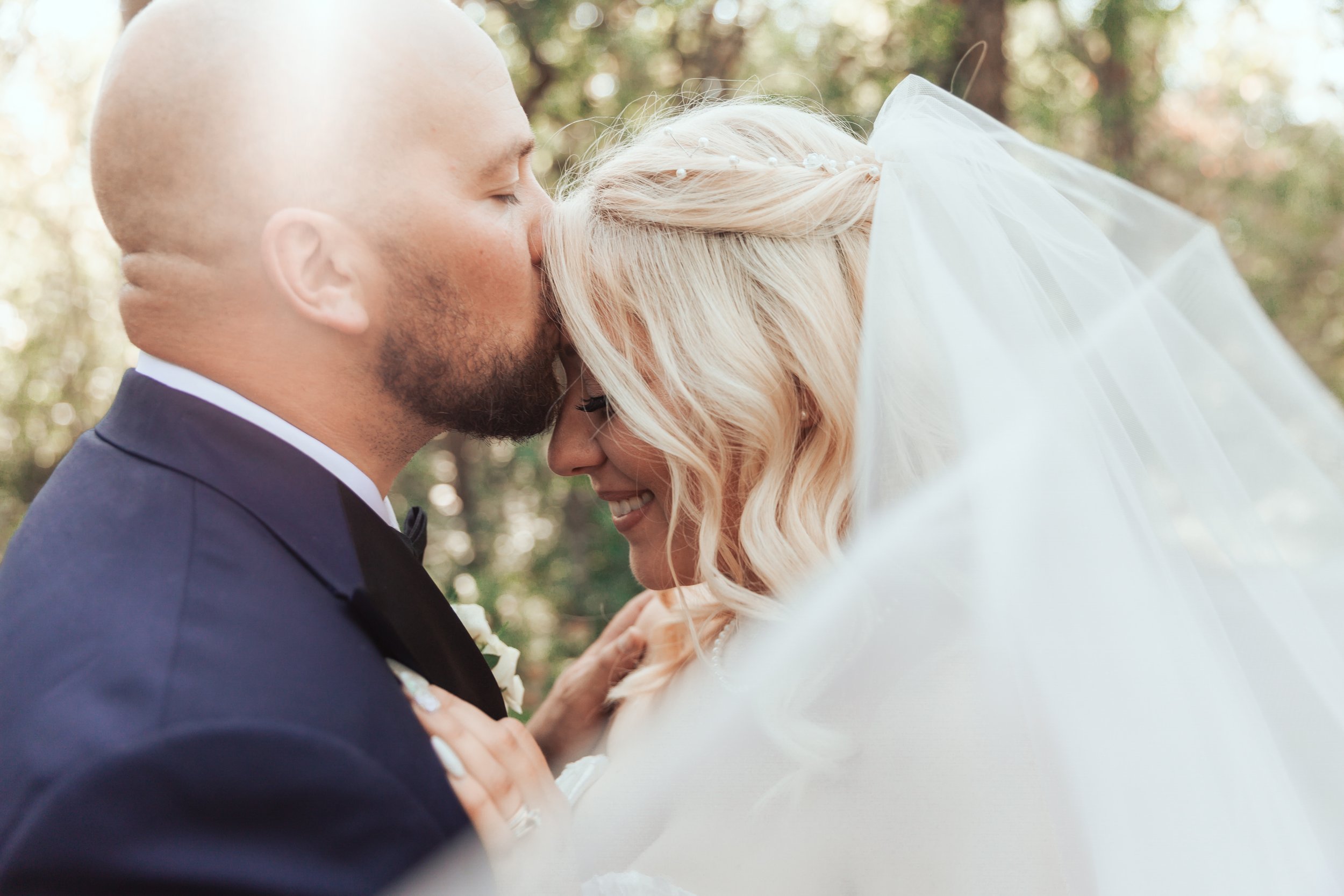 Groom kissing bride on the forehead outdoors, bride wearing veil