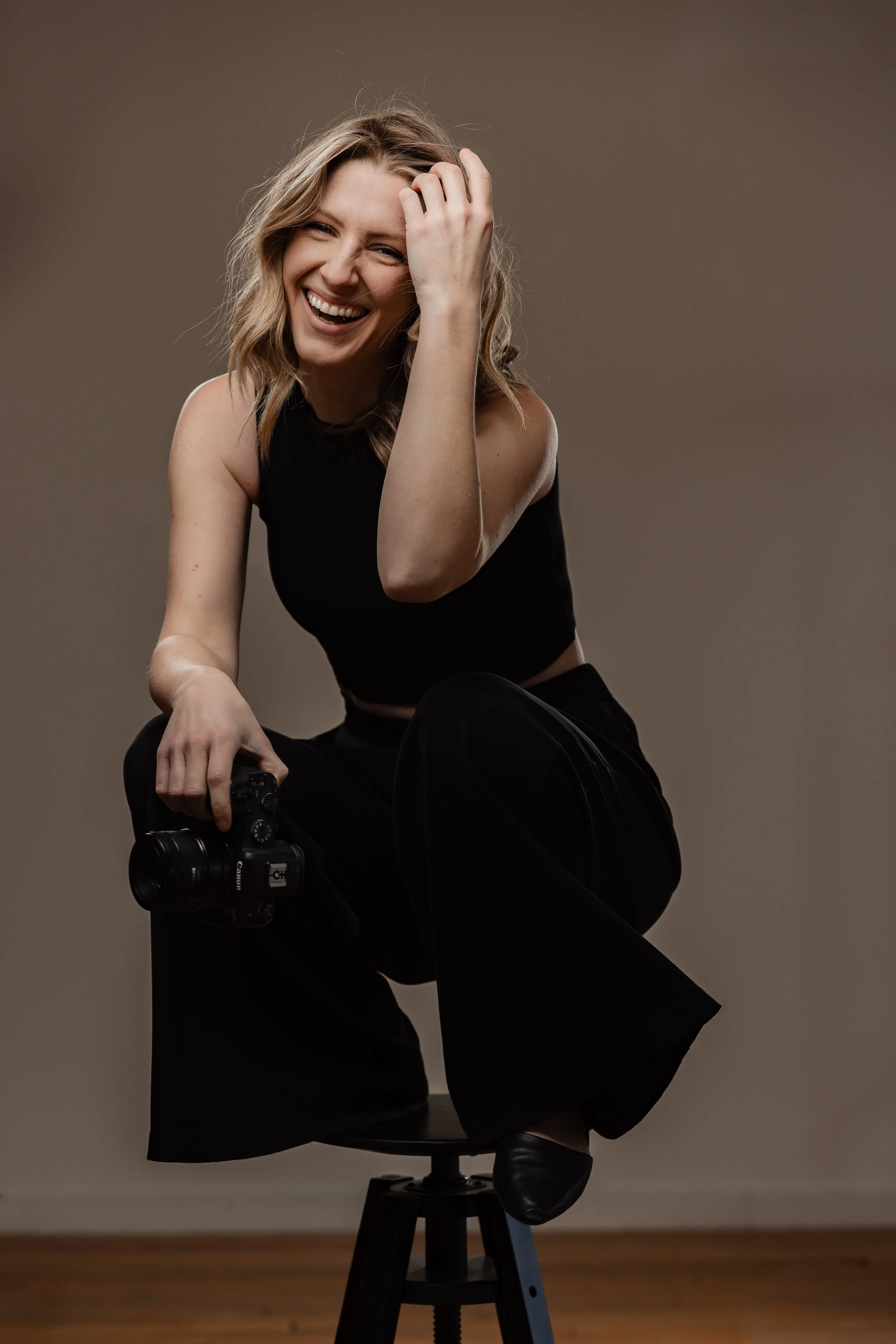 A young woman with blonde hair smiling, sitting on a stool, holding a camera in her right hand, wearing a black sleeveless top and black pants, with her left hand touching her hair.