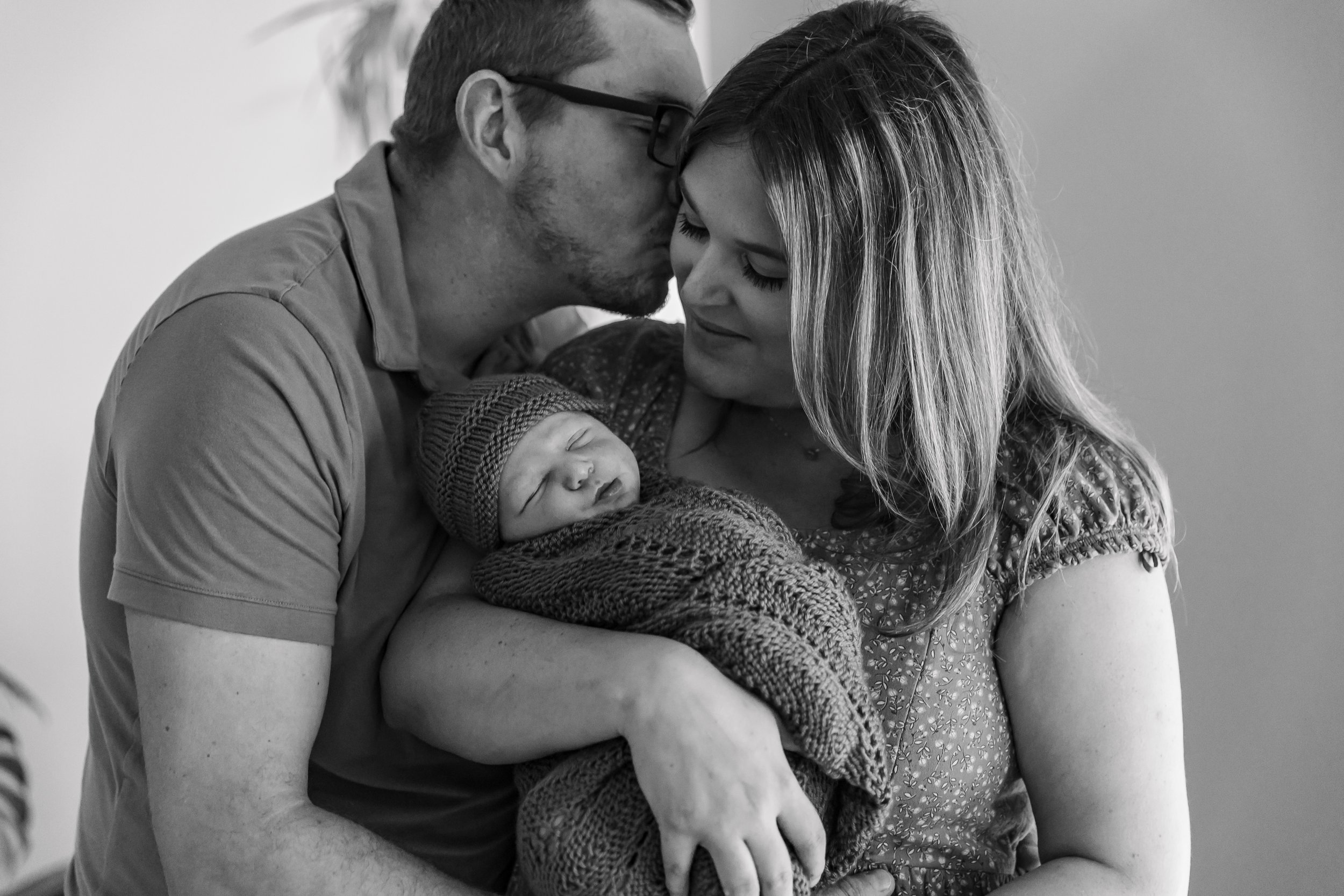 A black and white photo of a man kissing a woman's forehead while she holds a sleeping baby wrapped in a knitted blanket.