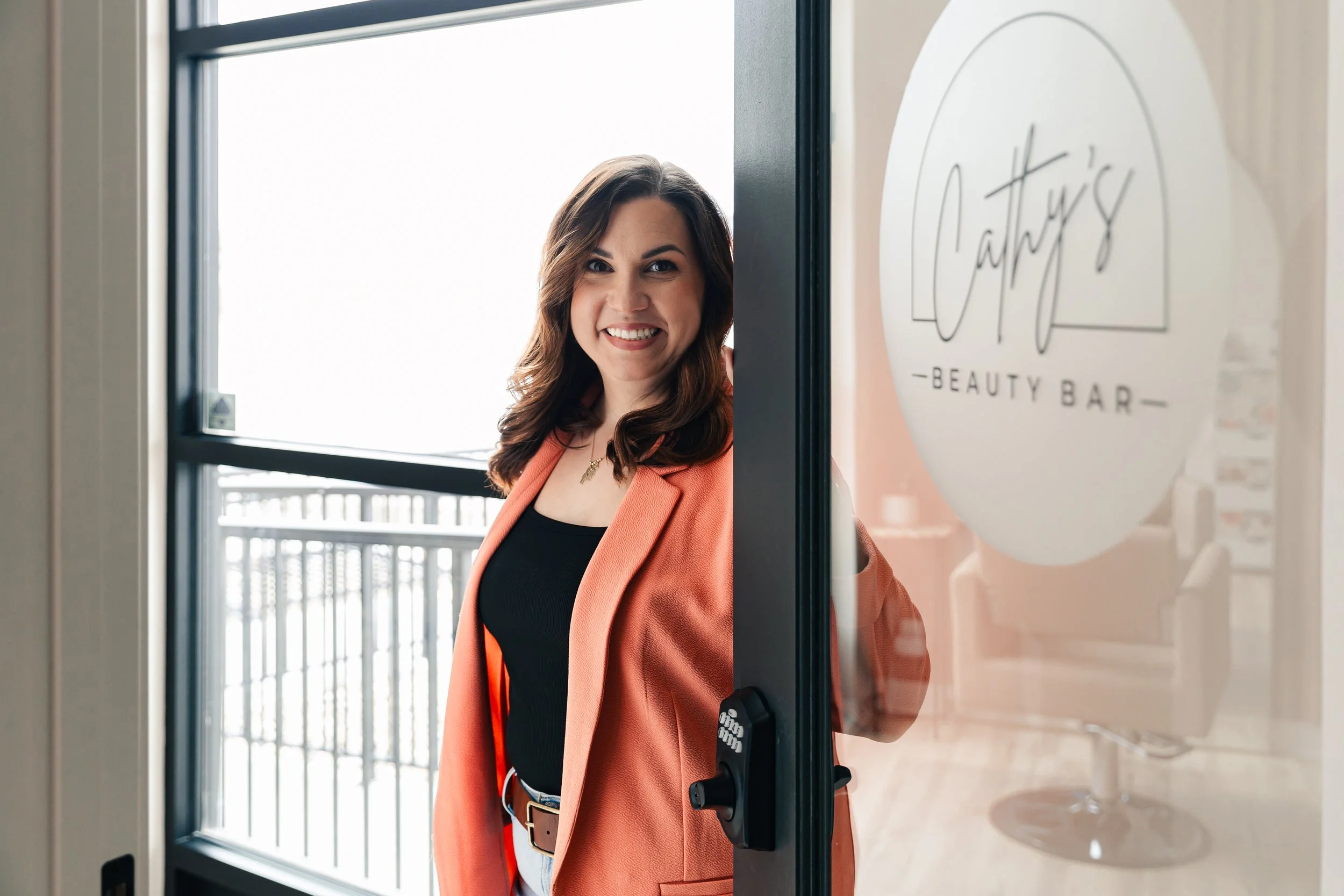 A woman with brown hair smiling and winking, standing at the door of a beauty bar called Cathy’s, wearing a coral blazer over a black top.