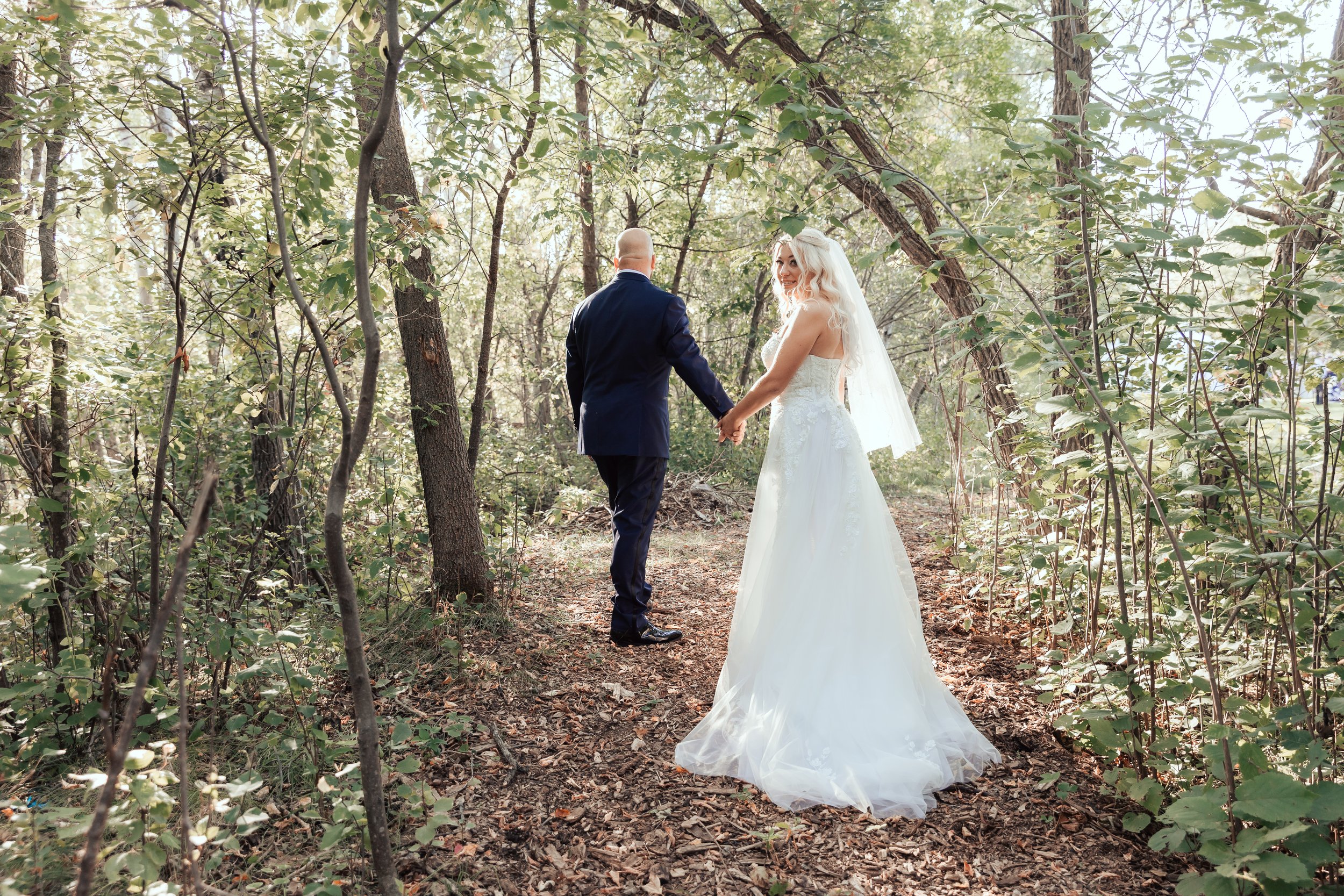 Bride and groom walking hand in hand through a forested path.