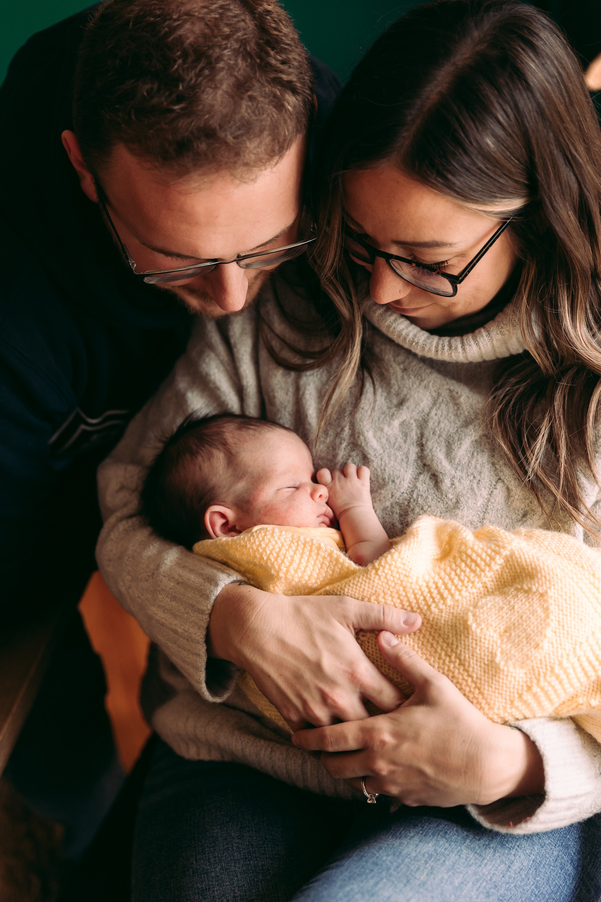 A couple gazes lovingly at a sleeping newborn wrapped in a yellow blanket.