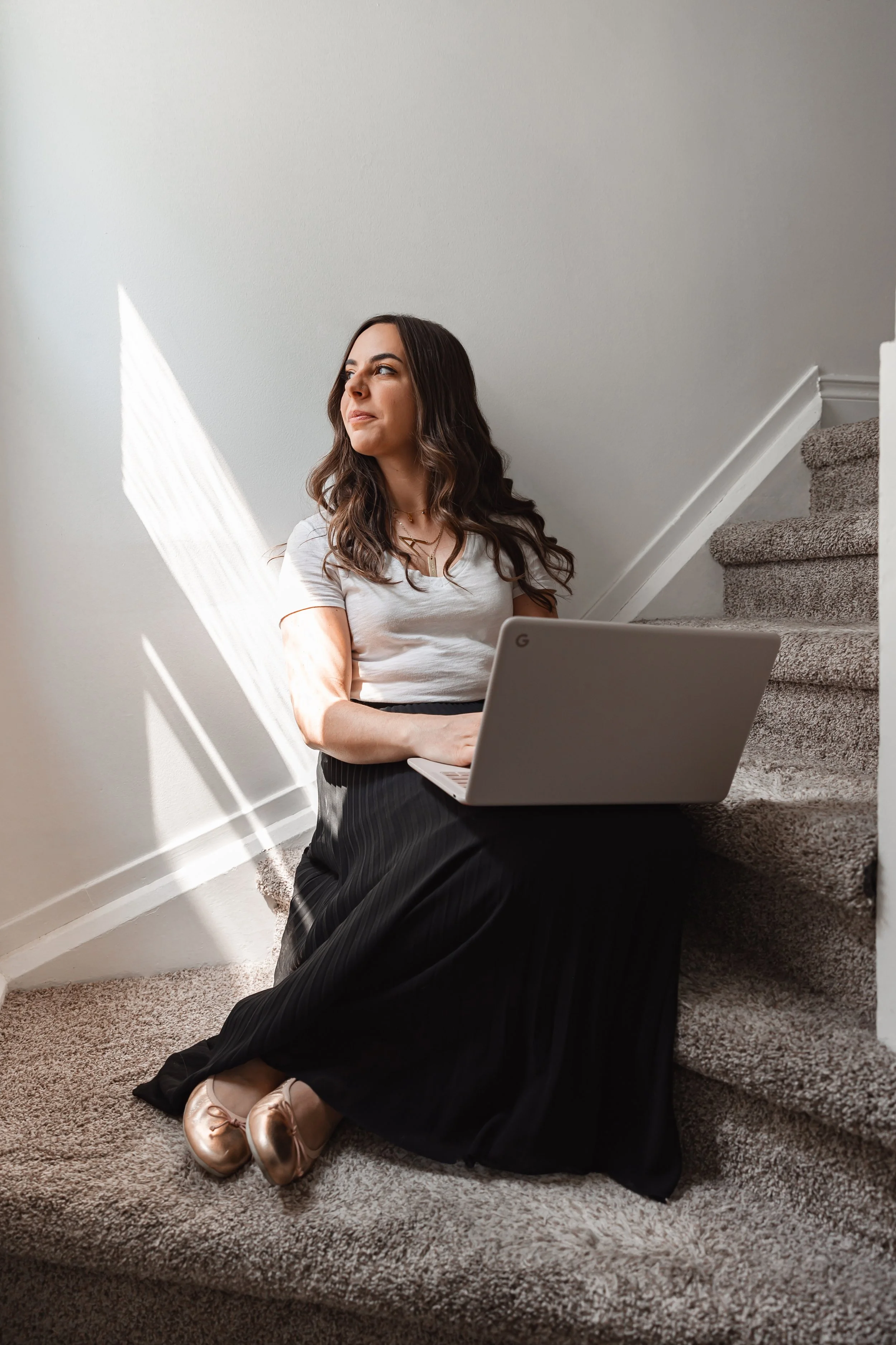 A woman with long, wavy brown hair sitting on carpeted stairs with a laptop in her lap, looking to her left while sunlight streams through a window, illuminating her face.