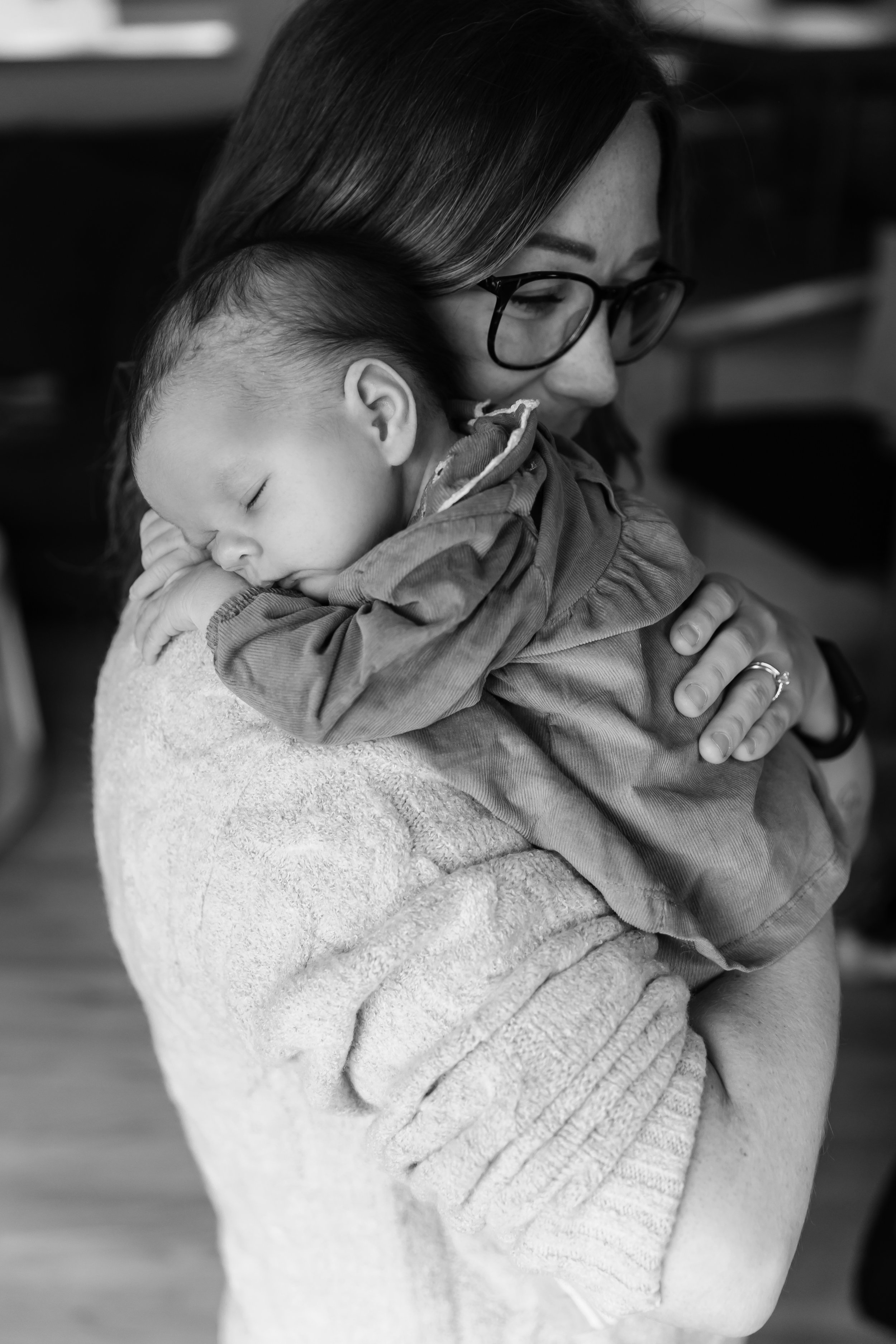 A black and white photo of a woman wearing glasses holding a sleeping baby on her shoulder.