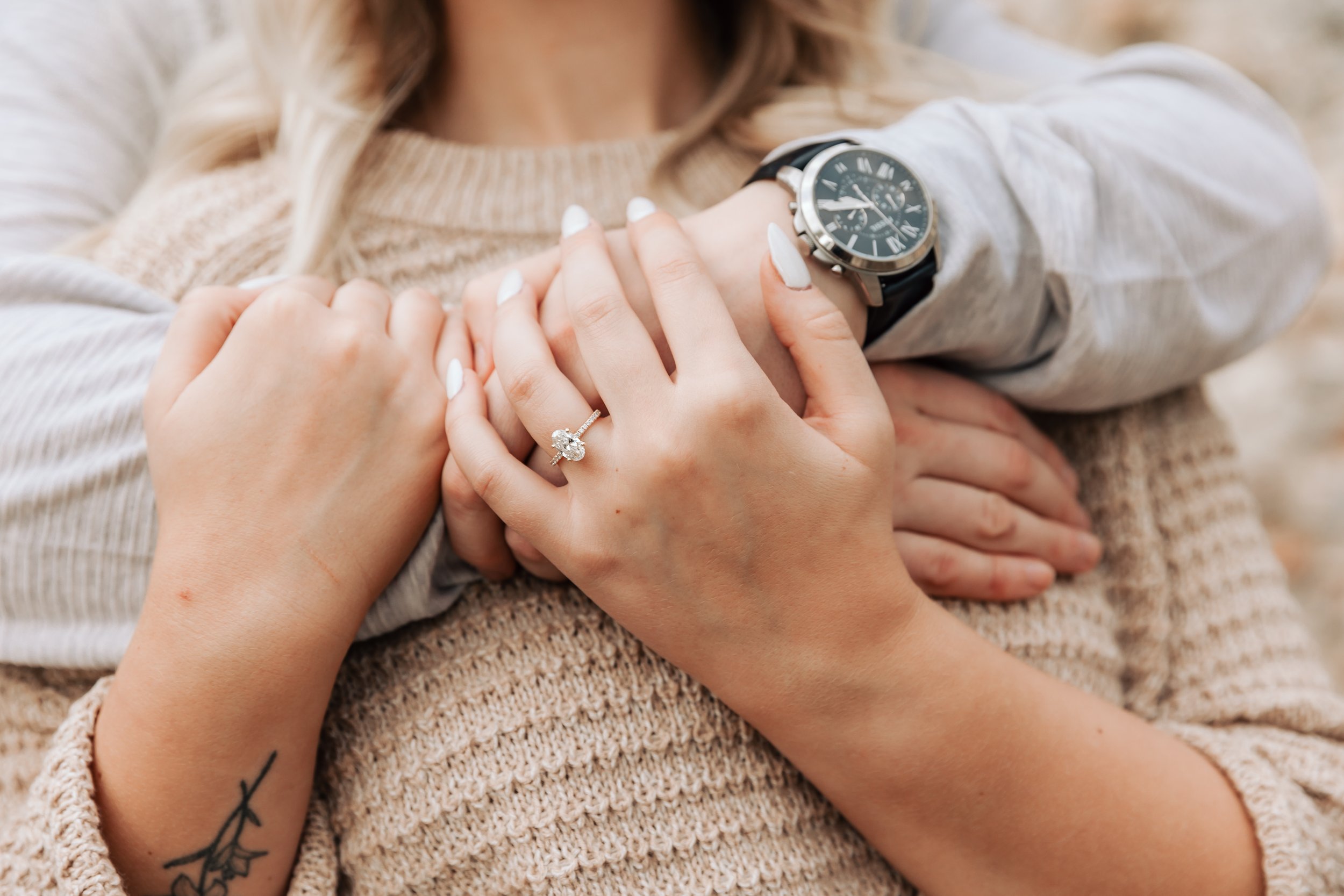 A close-up of intertwined hands, one with an engagement ring and another wearing a watch, against a person wearing a knit sweater.