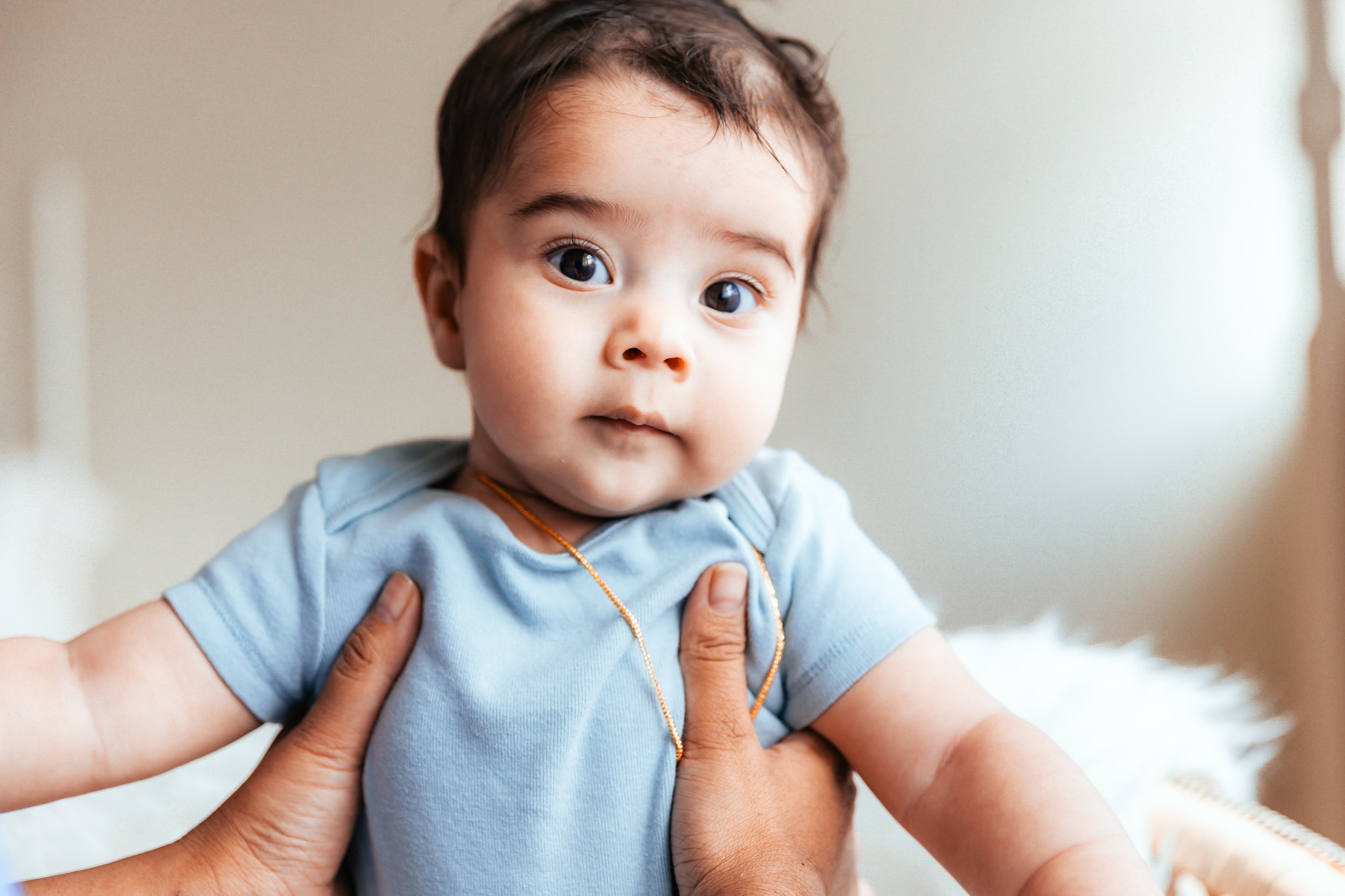 A baby being held by adult hands, wearing a blue onesie and a gold necklace, looking at the camera.
