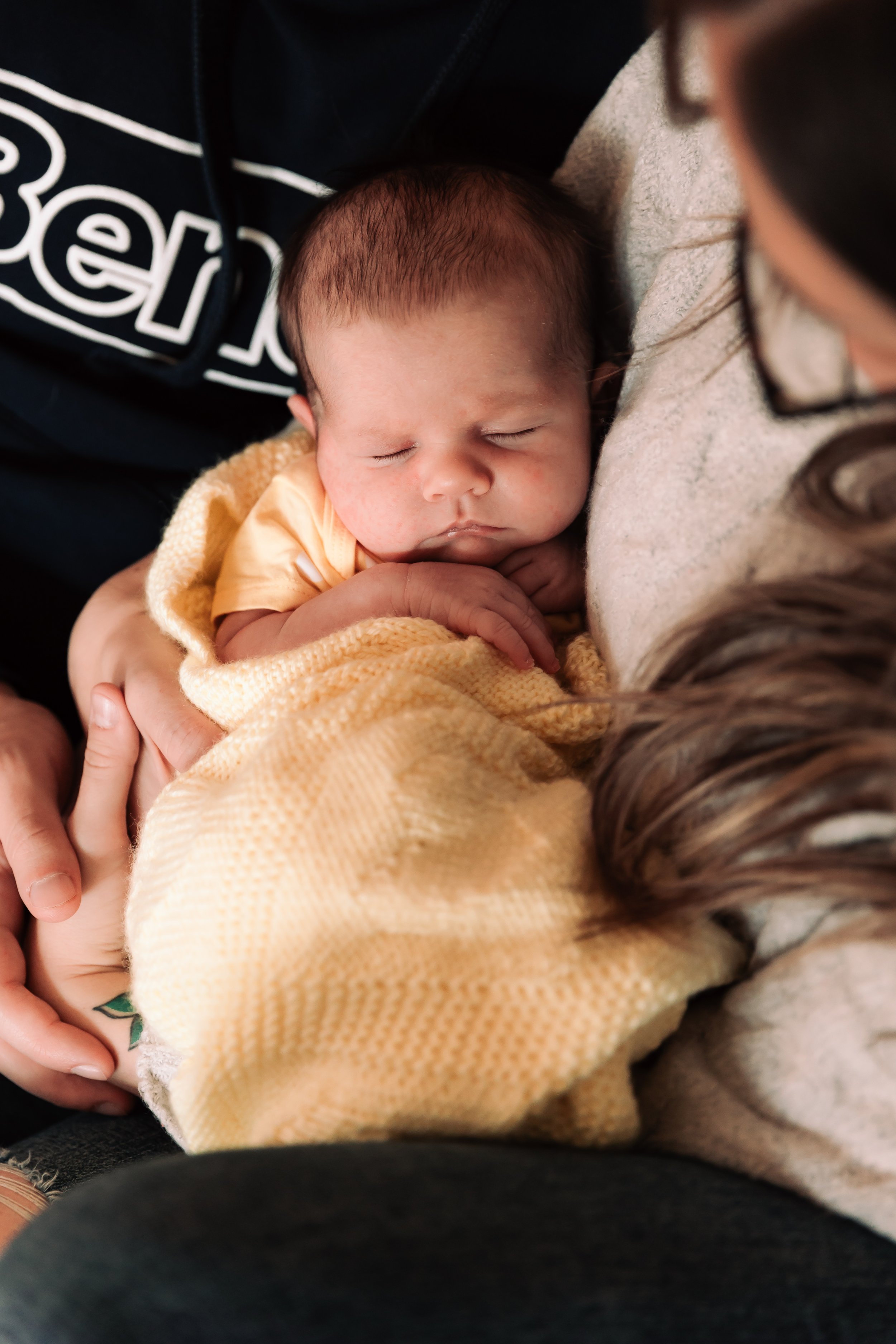 A sleeping baby wrapped in a yellow blanket being held by an adult, with part of a person's face visible.