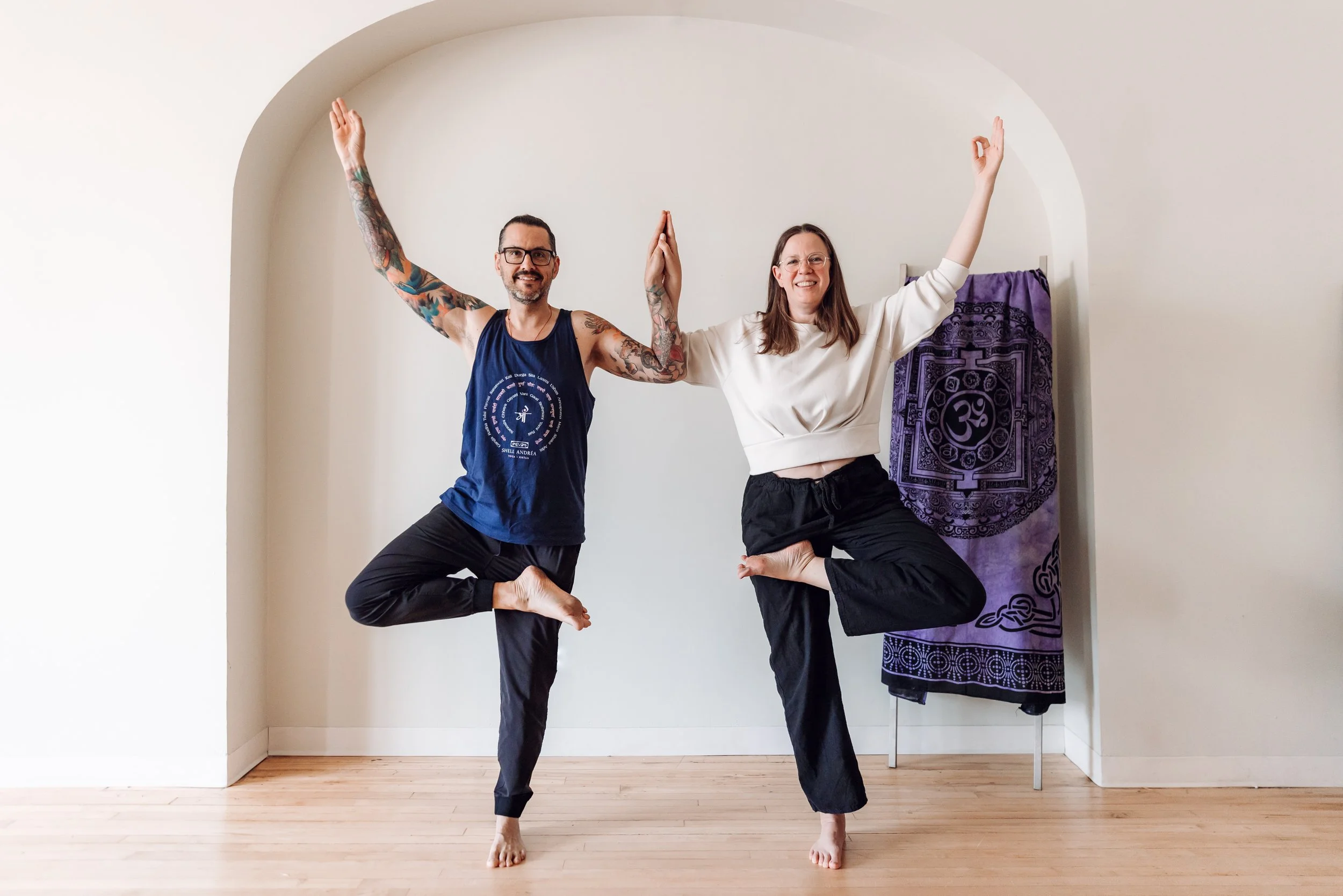 Two people practicing yoga during a branding session, standing on one leg with arms raised and hands in a mudra position, smiling in a well-lit room with a wooden floor and a purple curtain with spiritual symbols in the background.