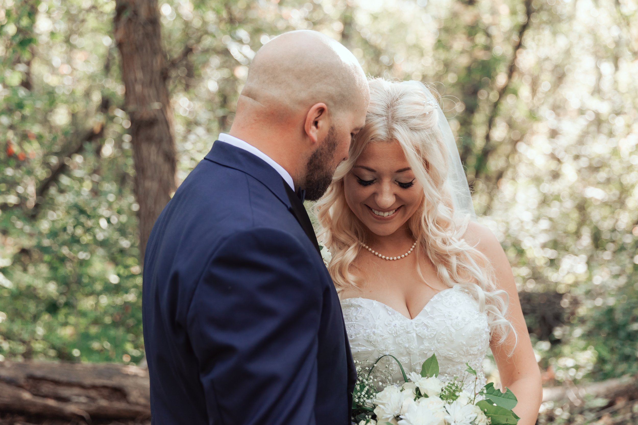 Bride and groom in wedding attire, smiling, with a bouquet of flowers, standing in a wooded area.