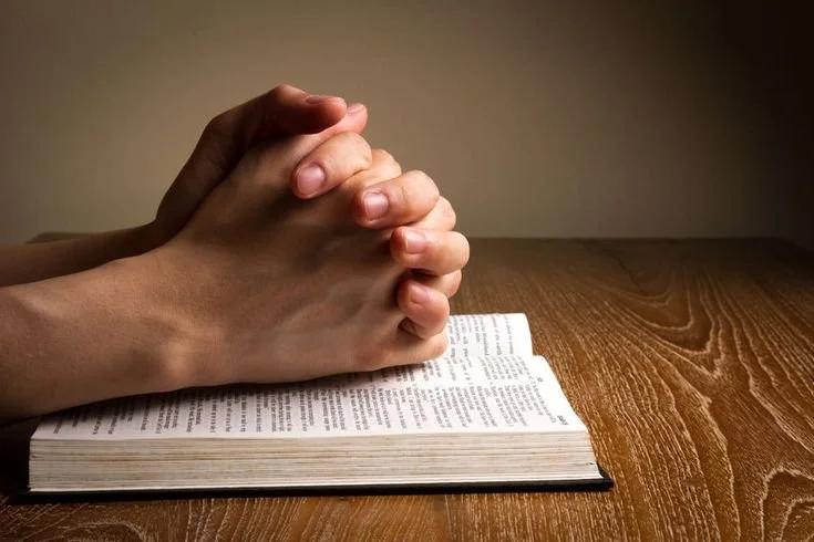 A person praying with hands clasped over an open book or bible on a wooden table.