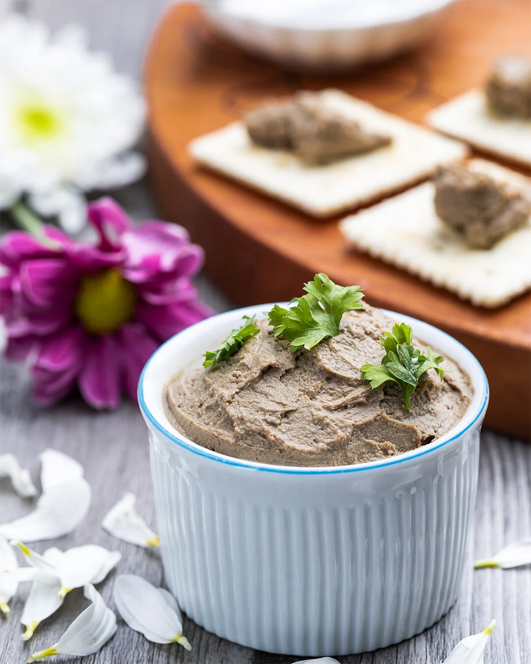 A white bowl filled with brown hummus garnished with fresh parsley, placed on a wooden surface with white flower petals around it. In the background, there are crackers topped with a meat spread and a purple flower on the left.