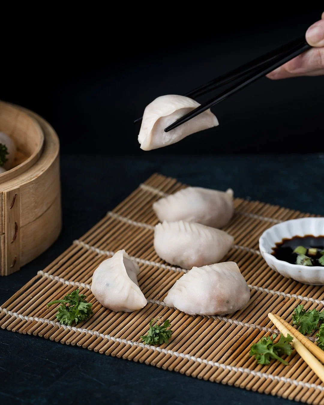 Chinese dumplings on a bamboo mat, with soy sauce in a white dish and garnished with parsley, with chopsticks picking up one dumpling.