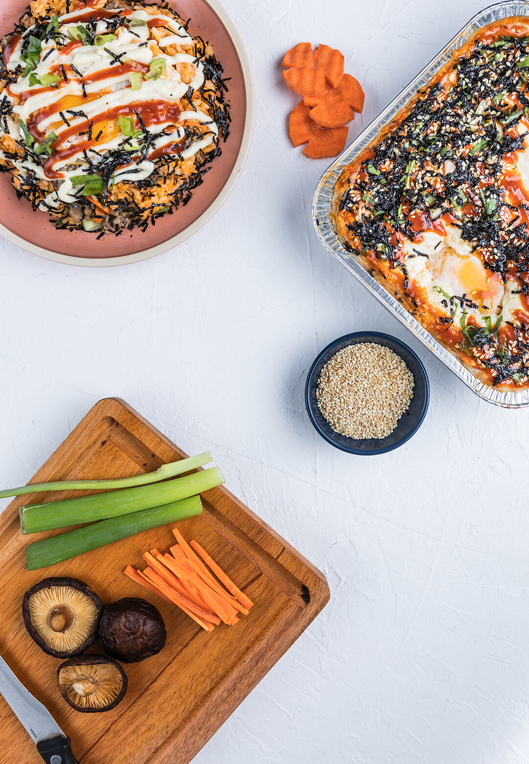 A top-down view of a table with an uncooked vegetable bibimbap and a baked casserole with egg, rice, and black seaweed strips, alongside a small bowl of sesame seeds. There is also a wooden cutting board with sliced carrots, green onions, and shiitak