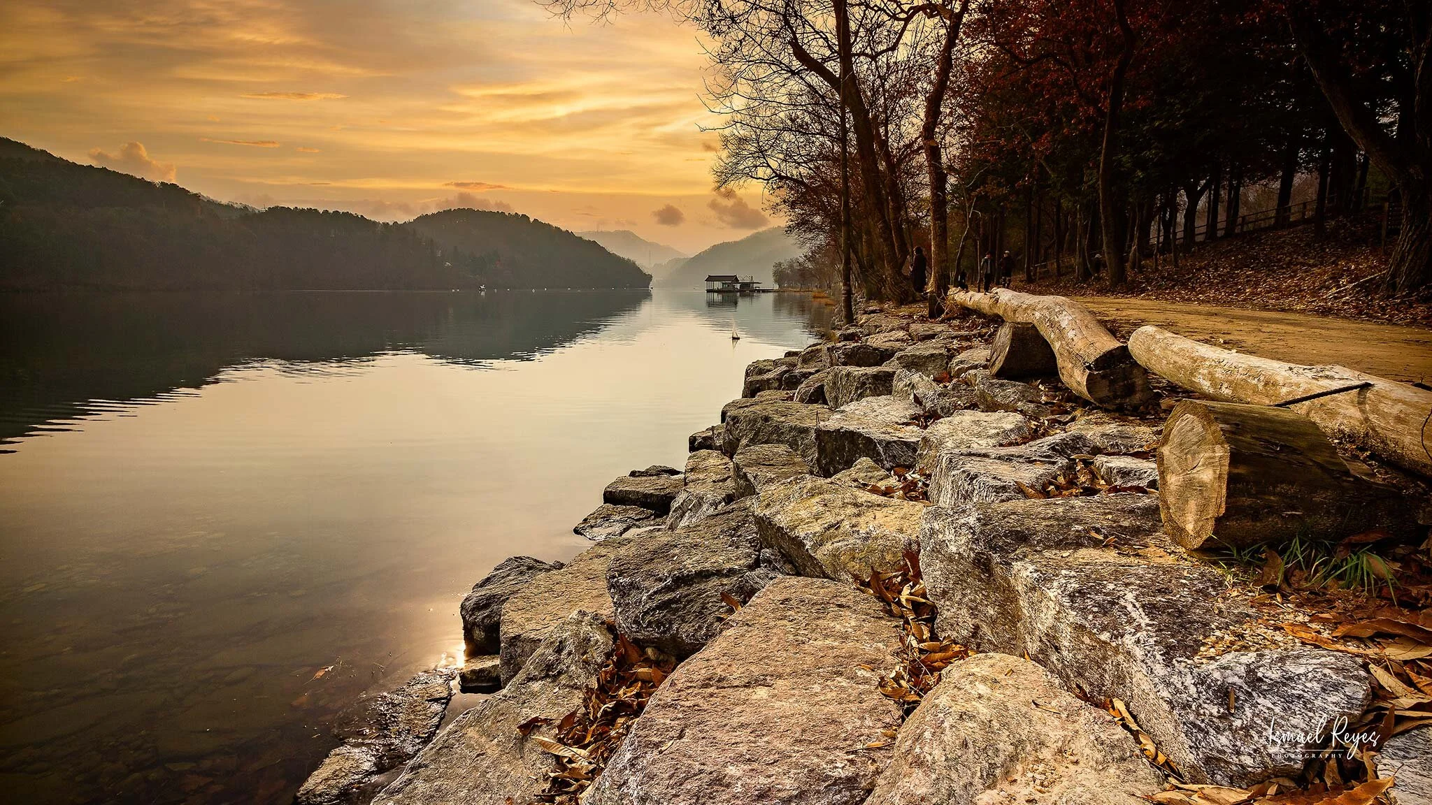 Sunset over a calm lake with hilly mountains in the background, a rocky shoreline, and trees lining the path. There are some people walking on the path, and a small house on stilts above the water in the distance.