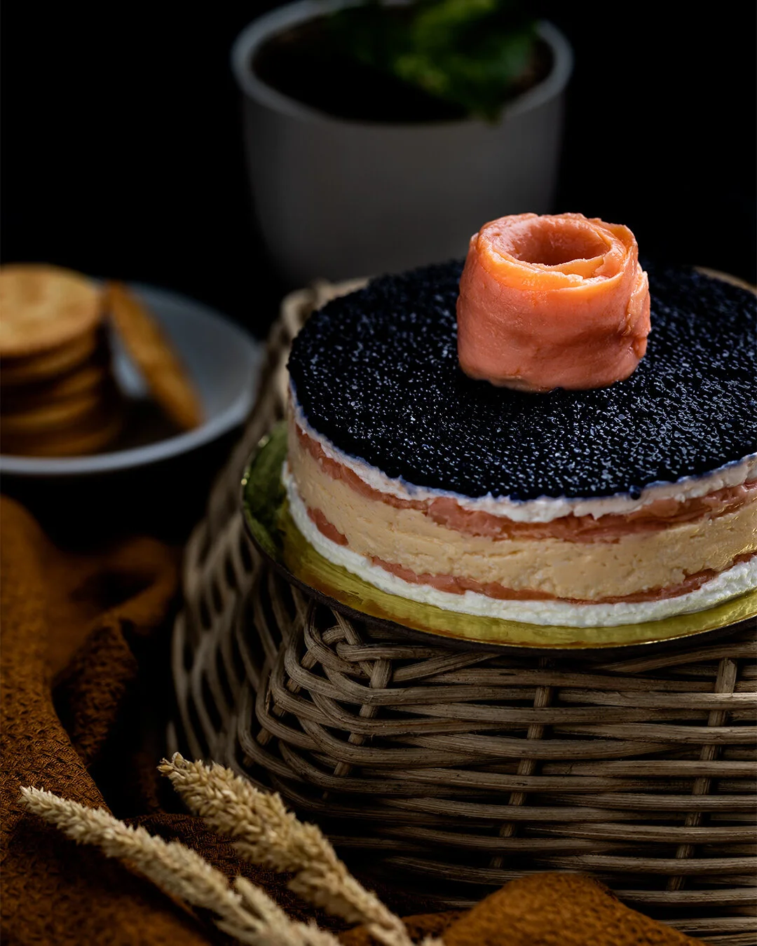 Cheesecake topped with black caviar and a rolled slice of ham on top, with a cup of green tea or coffee and cookies in the background