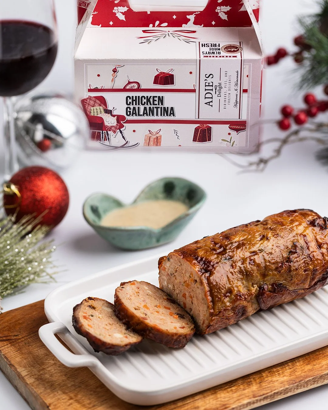 A baked chicken loaf on a white dish with a few slices cut, placed on a wooden cutting board. In the background, there is a holiday-themed box labeled "Chicken Galantina," along with a glass of red wine, a small green sauce dish, and holiday decorati