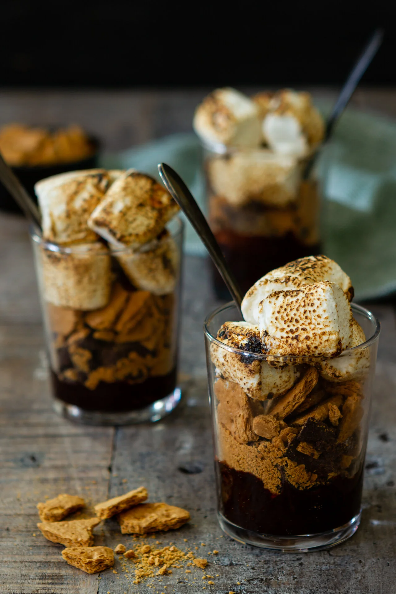 Three glasses of s'mores-inspired dessert with chunks of graham crackers, chocolate, and toasted marshmallows, on a rustic wooden surface.
