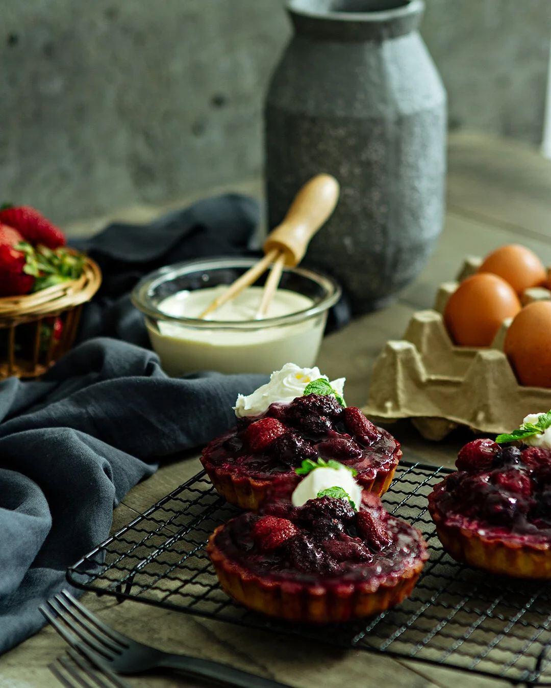 Three cherry berry tarts decorated with whipped cream and mint leaves sit on a wire rack. In the background, there is a bowl of cream, a basket of strawberries, a carton of eggs, and a large ceramic vase, all on a rustic wooden table with a dark clot